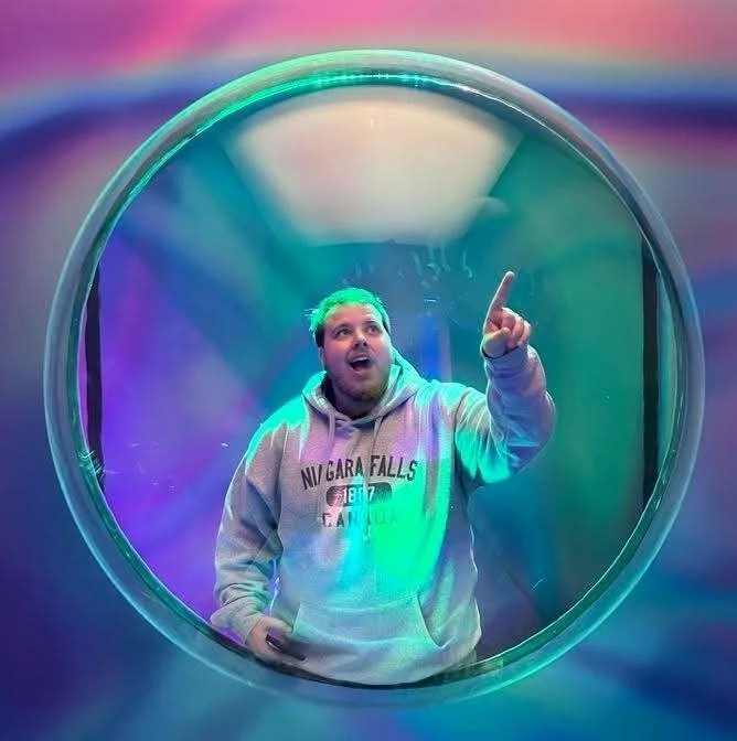 A man wearing a gray hoodie, taking a selfie inside a large, transparent, circular structure with colorful, neon lighting.