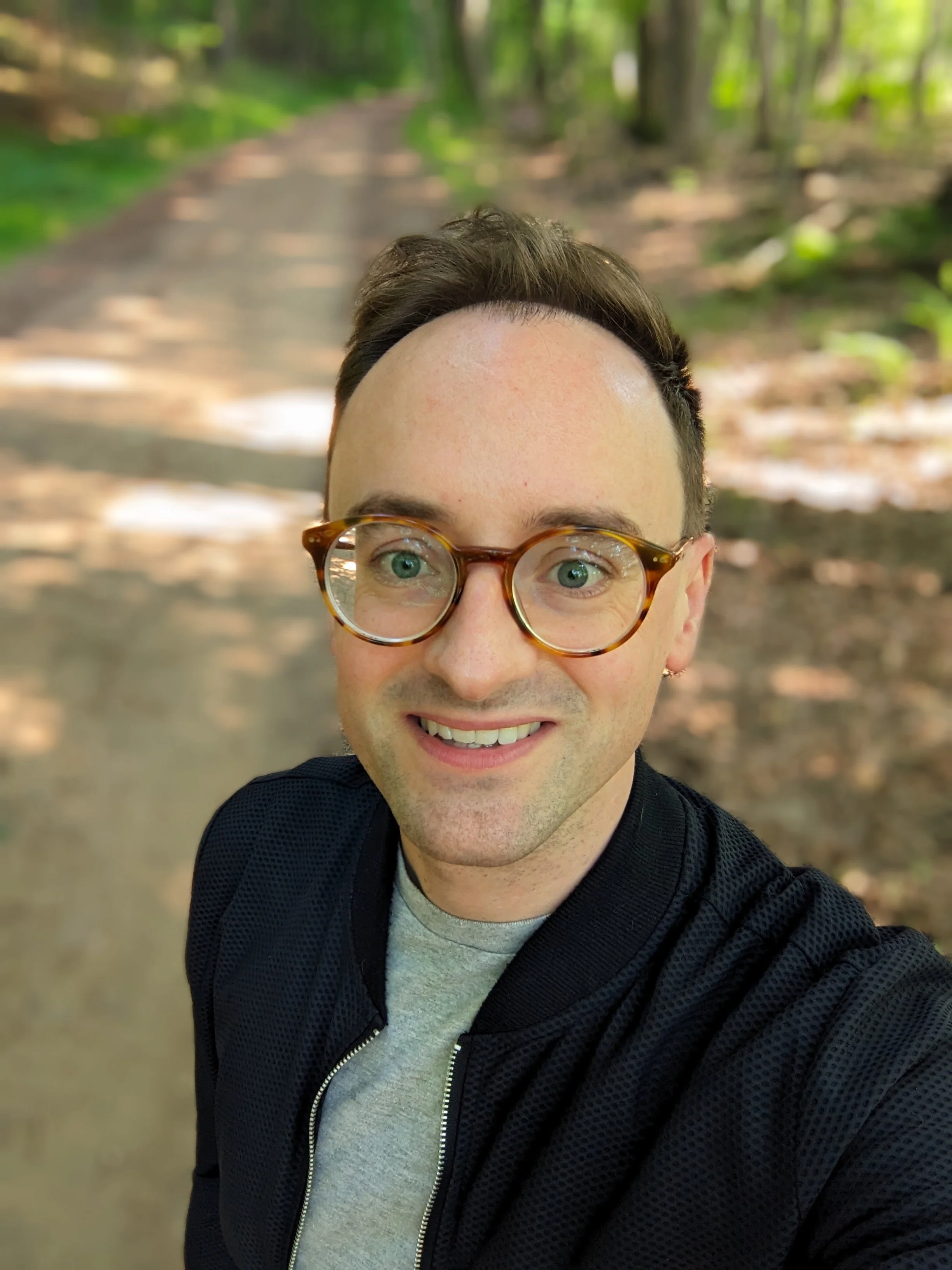 A man with glasses smiling outdoors on a forest trail. The background shows trees and greenery.