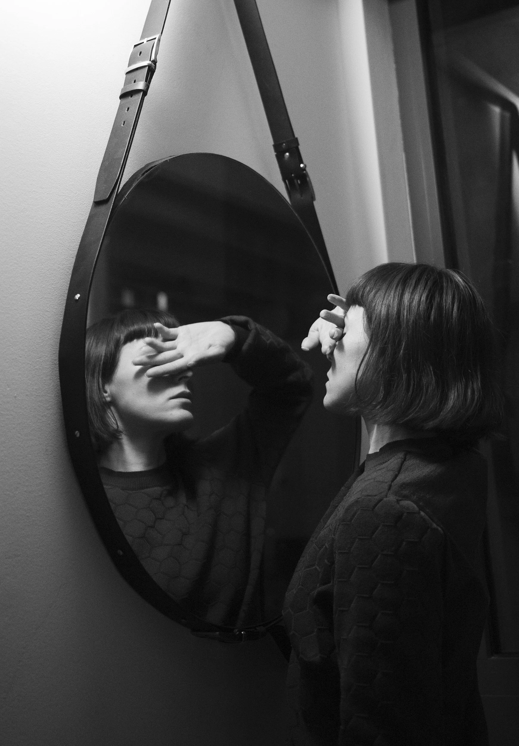 A young woman with shoulder-length hair looking at her reflection in a round mirror, with her hand covering part of her face, in an indoor setting.