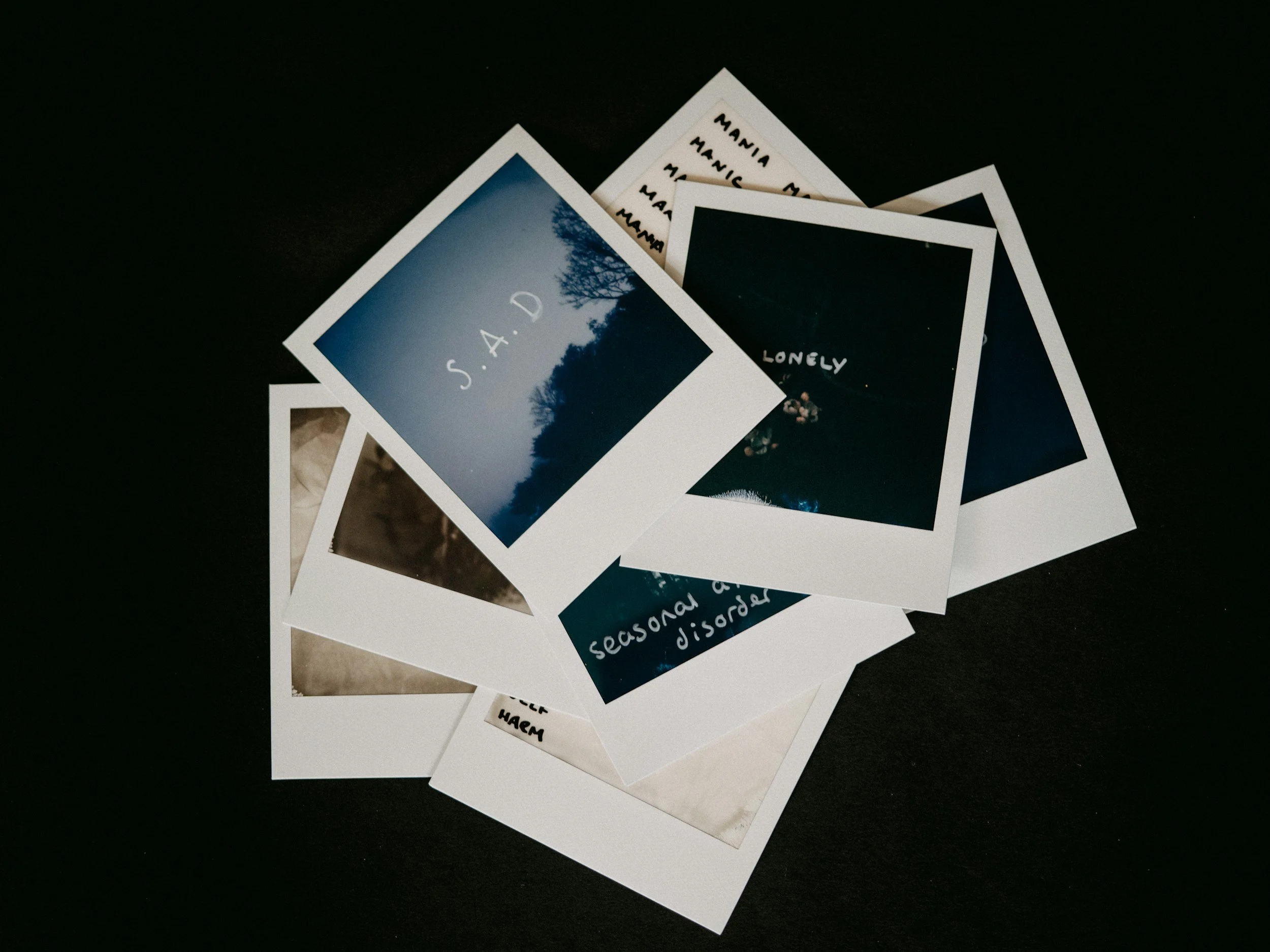 A collection of printed photographs, with one showing a tree silhouette against a blue sky and the text 'S. A. D.' on top. Other photos contain text related to emotions such as 'lonely' and 'sad,' and some have handwritten labels.