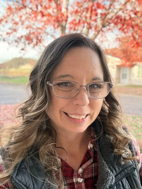 A woman with glasses smiling outdoors during autumn, with red and orange leaves in the background.