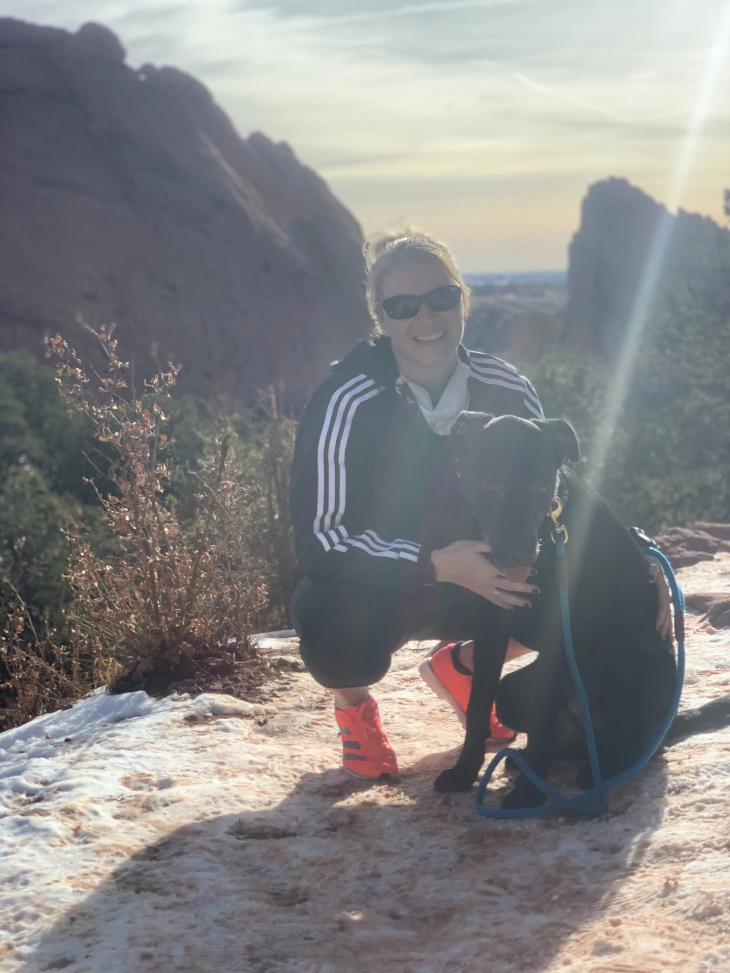 A woman crouching on snowy ground in a mountainous outdoor setting, smiling at the camera, wearing sunglasses, a striped jacket, bright red rain boots, and holding a black dog. There are mountains and a partly cloudy sky in the background, with sunlight creating a lens flare.