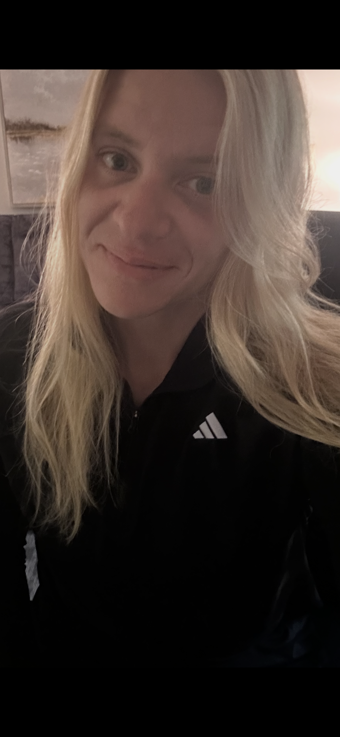 A woman with long blonde hair and fair skin taking a selfie, wearing a black jacket with a small white logo, in a room with dim lighting.