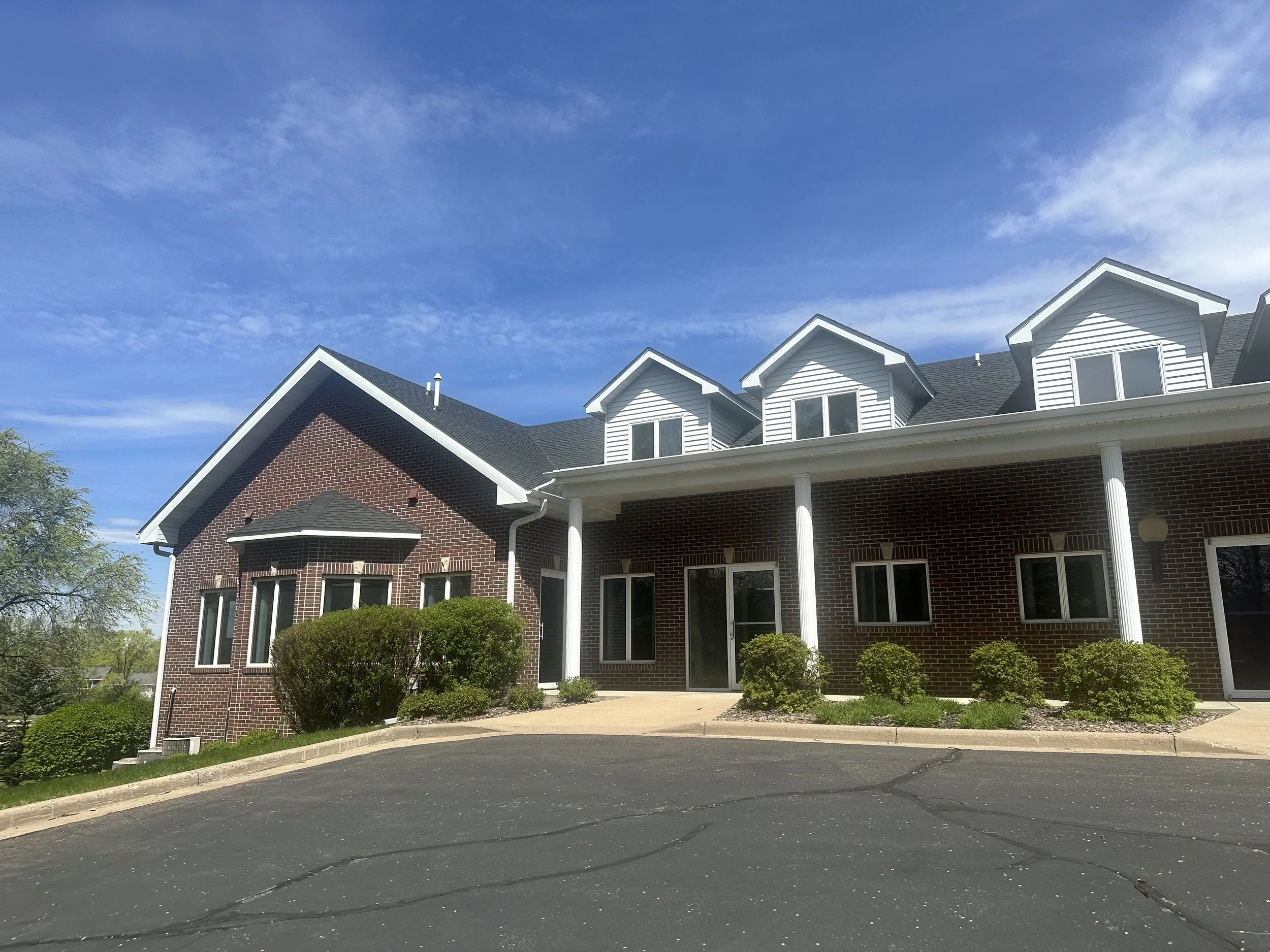 A house with a brick lower level and a white upper level with three gabled dormers, black roof, and white columns at the porch, under a blue sky with some clouds.