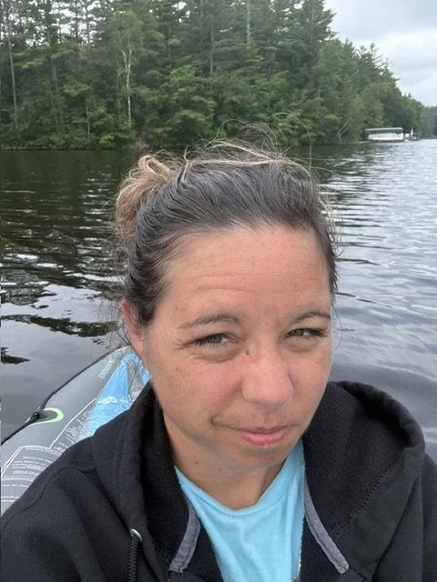 A woman with brown hair in a bun taking a selfie on a boat or kayak on a river, with trees along the shoreline in the background under a cloudy sky.