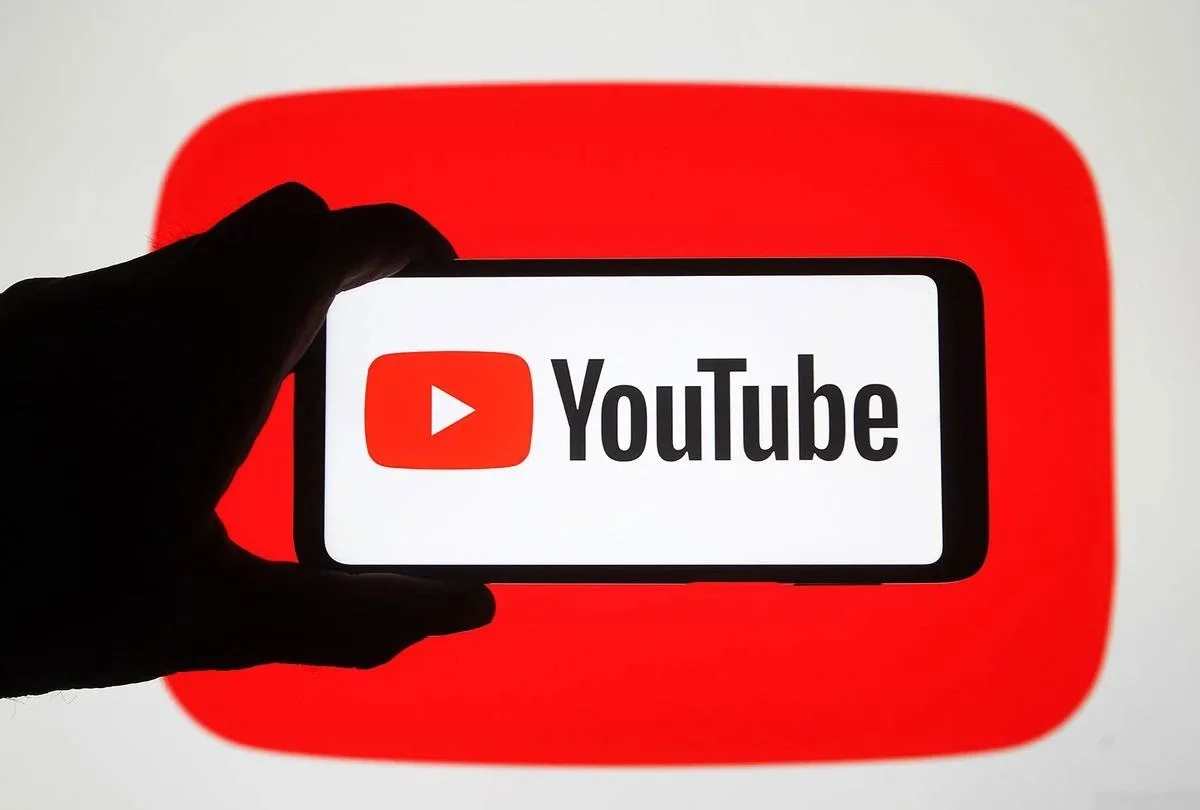Hand holding a smartphone displaying the YouTube logo in front of a large YouTube icon background.