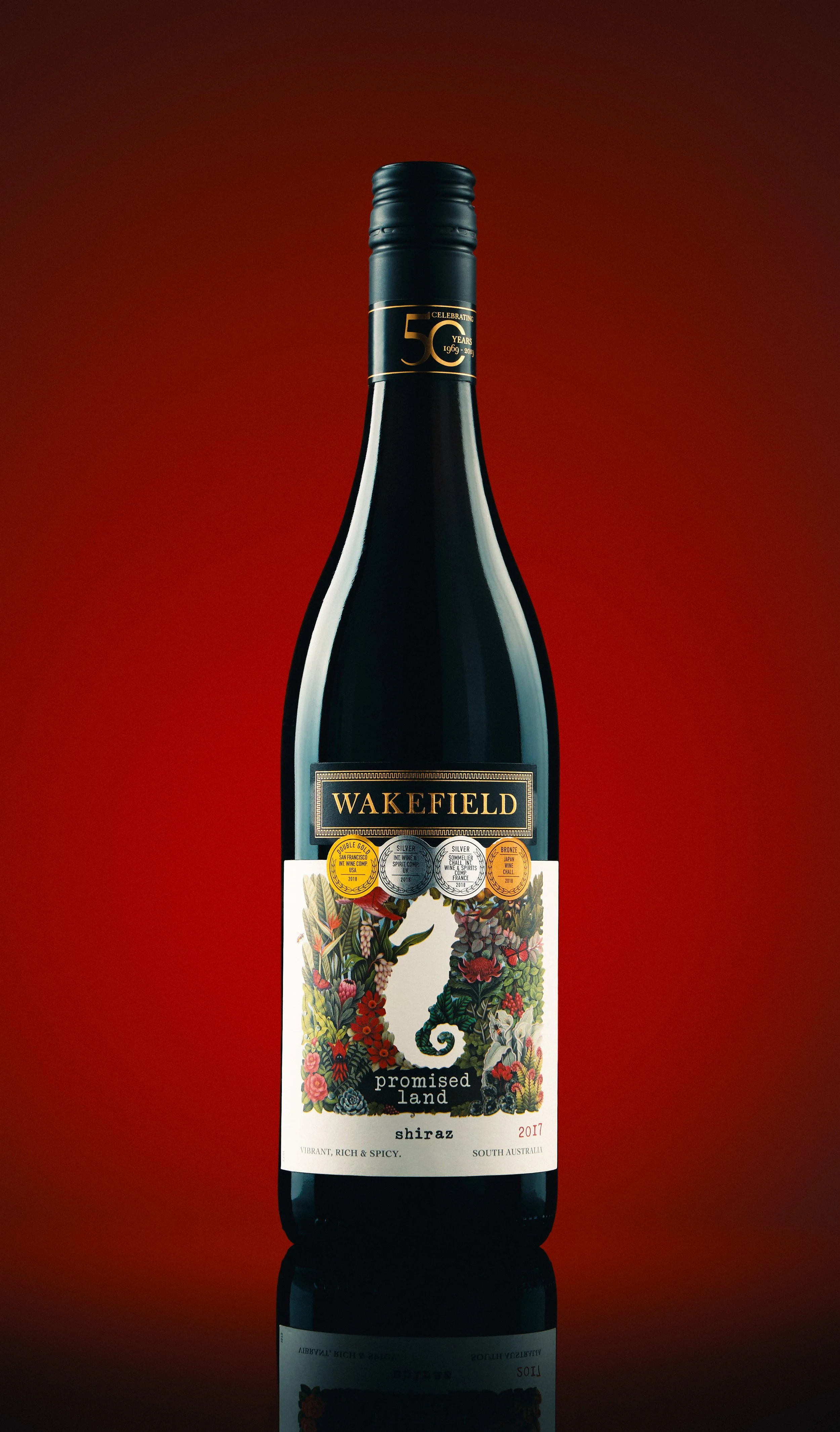 A bottle of Wakefield Promised Land Shiraz wine from South Australia, 2017, with a black cap and a label depicting colorful plants and flowers, set against a red background.