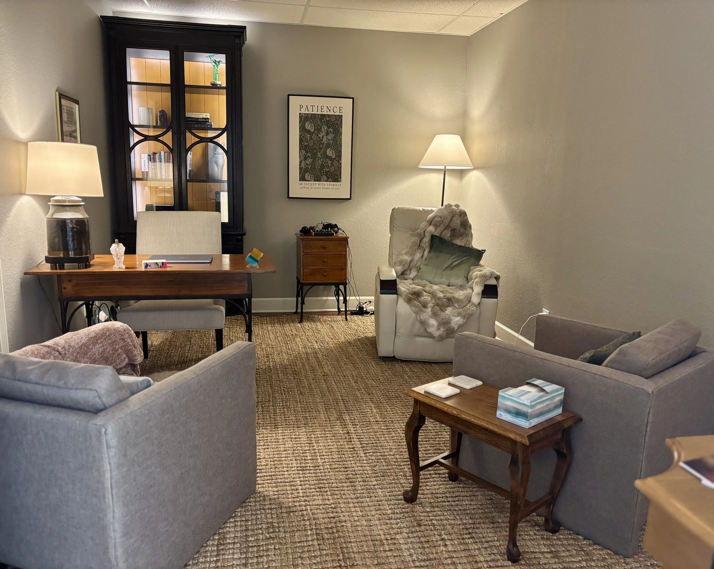 A cozy living room corner with a grey sofa, an armchair with a fur throw, a wooden side table with tissue box, a wooden desk with a lamp and small items, a black display cabinet with glasses and decor, and walls decorated with framed art and a poster that says 'Patience'.