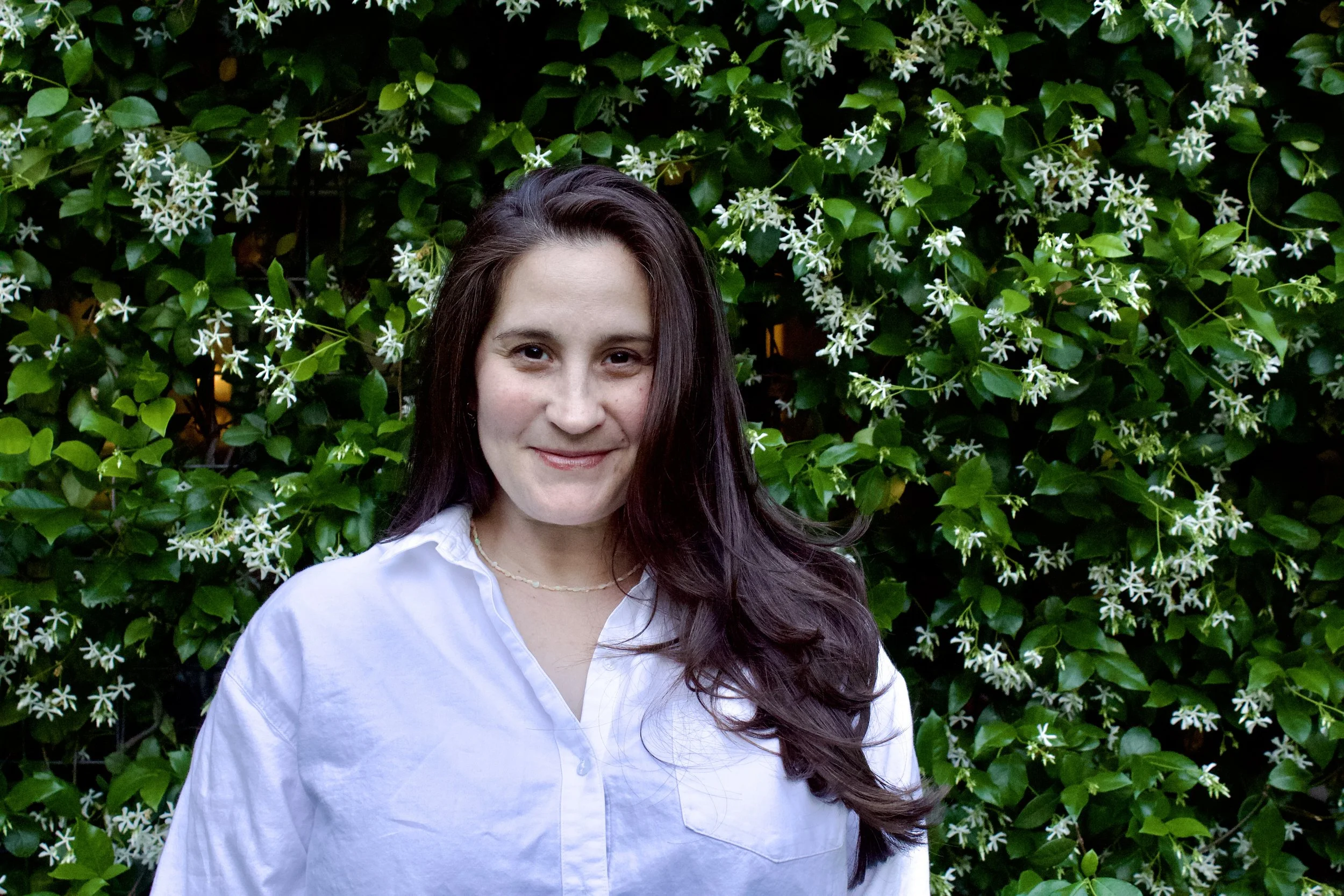 A woman with long brown hair wearing a white shirt and a necklace, standing in front of a green leafy bush with white flowers.