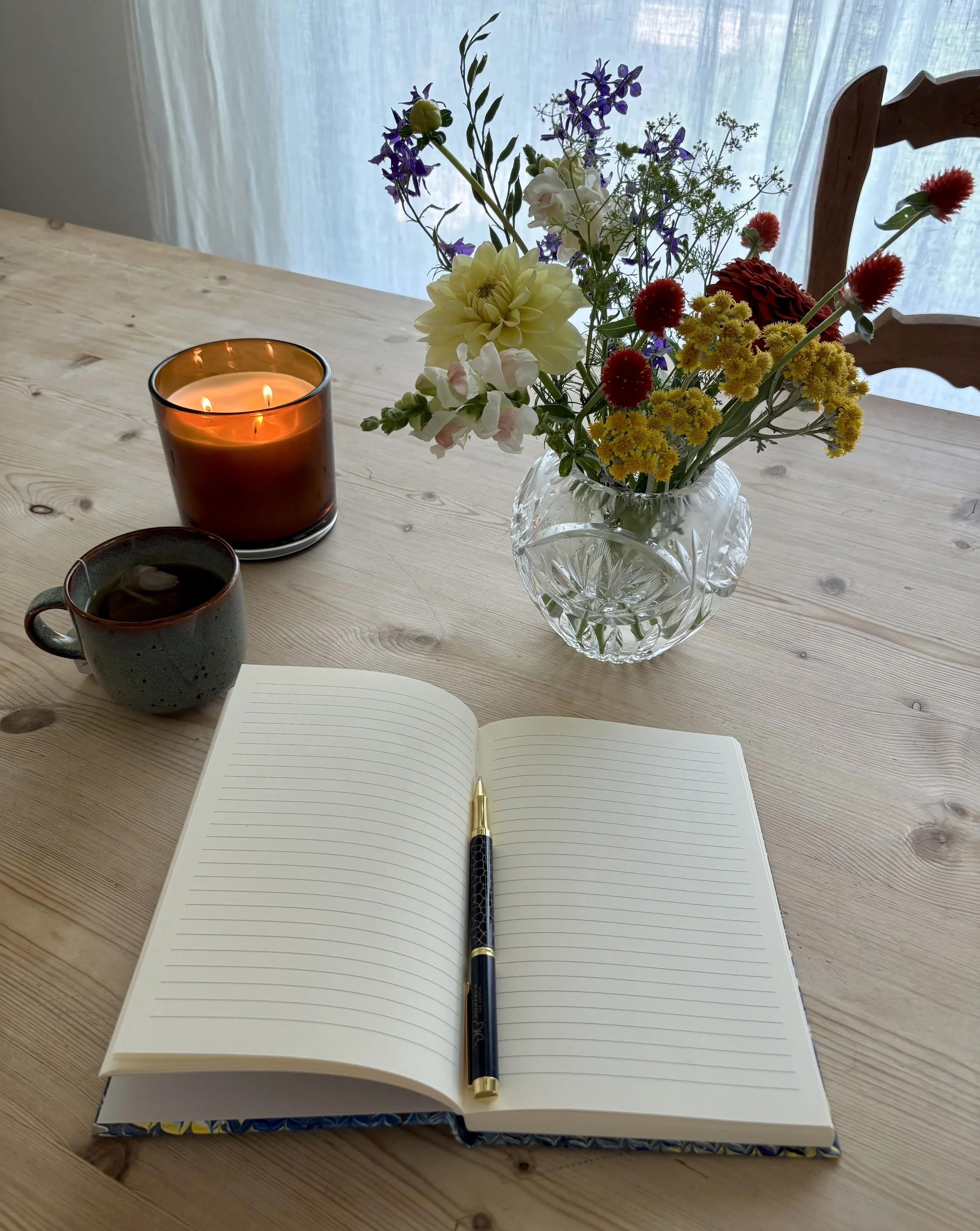 Open blank lined notebook with pen on top, placed on a wooden table, with a bouquet of colorful flowers in a glass vase and a lit candle also on the table