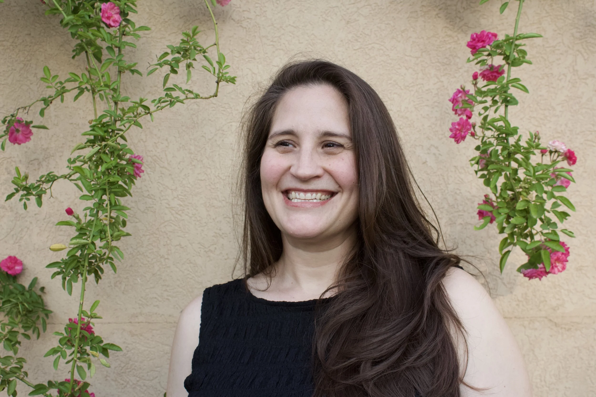 A woman with long brown hair smiling in front of a beige stucco wall with pink flowering plants on either side.