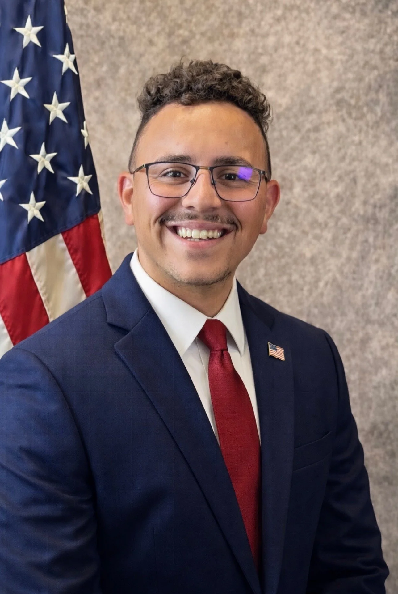 Keegan Hall smiling, wearing glasses, a navy suit, white shirt, and red tie, standing in front of an American flag.