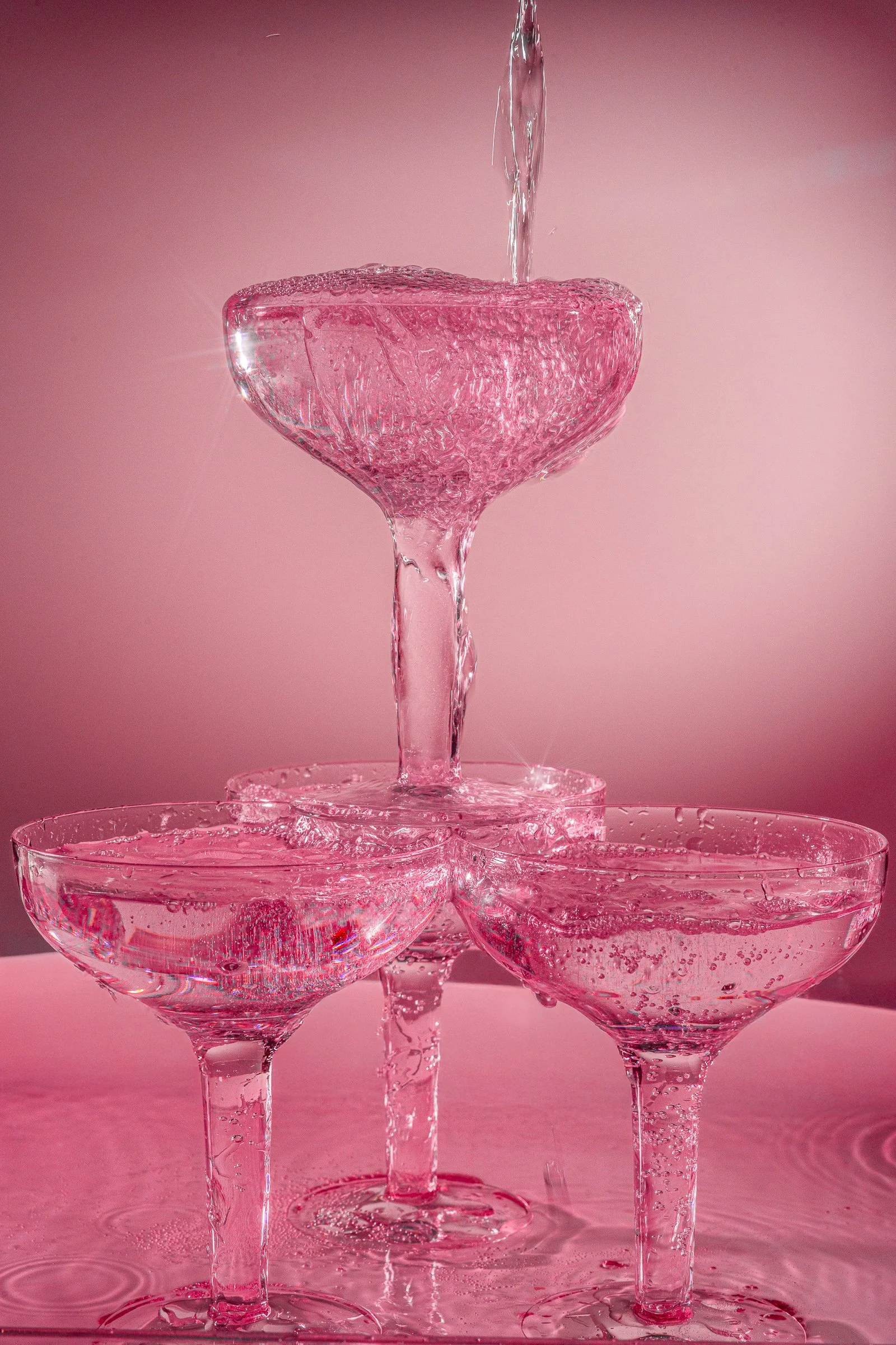 A pyramid of five pink glass cocktail glasses filled with water, with water pouring into the top glass, creating splashes and bubbles.