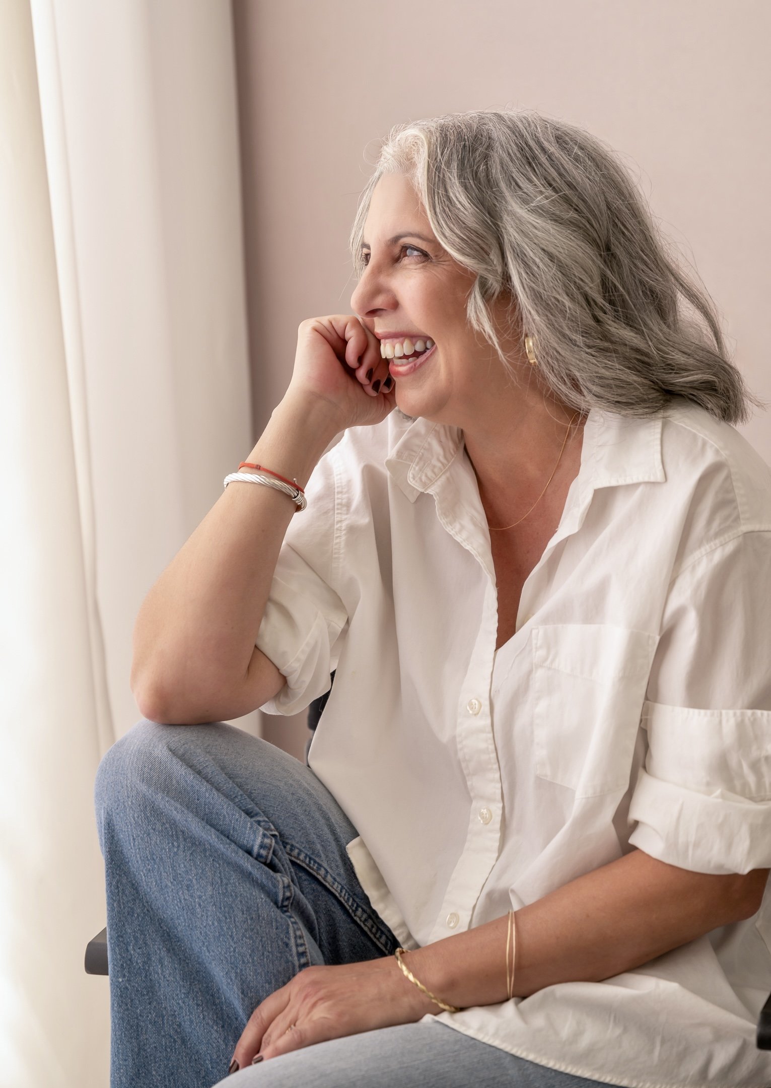 A cheerful middle-aged woman with gray hair, wearing a white button-down shirt and jeans, sitting near a window with a smile on her face.