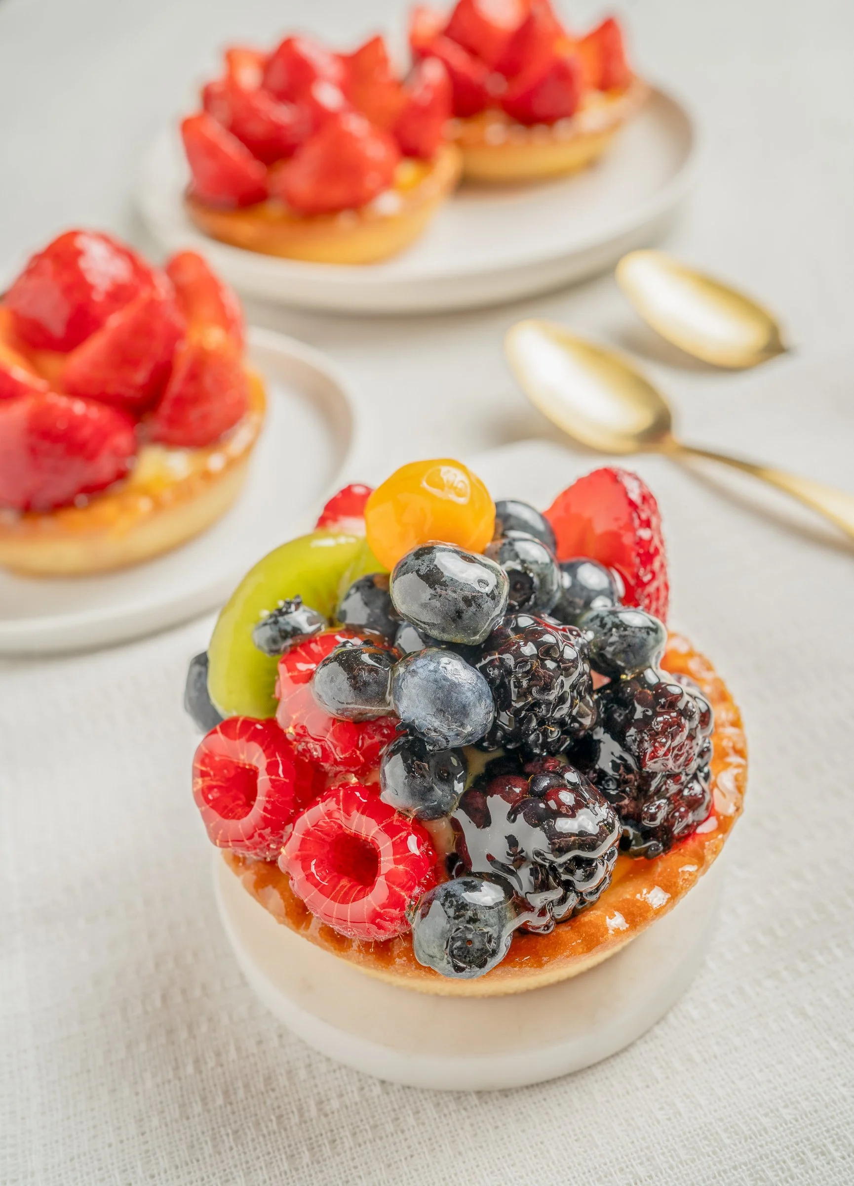 Close-up of a fruit tart topped with mixed berries, including blueberries, raspberries, strawberries, and slices of kiwi and orange, on a white plate with a white background.
