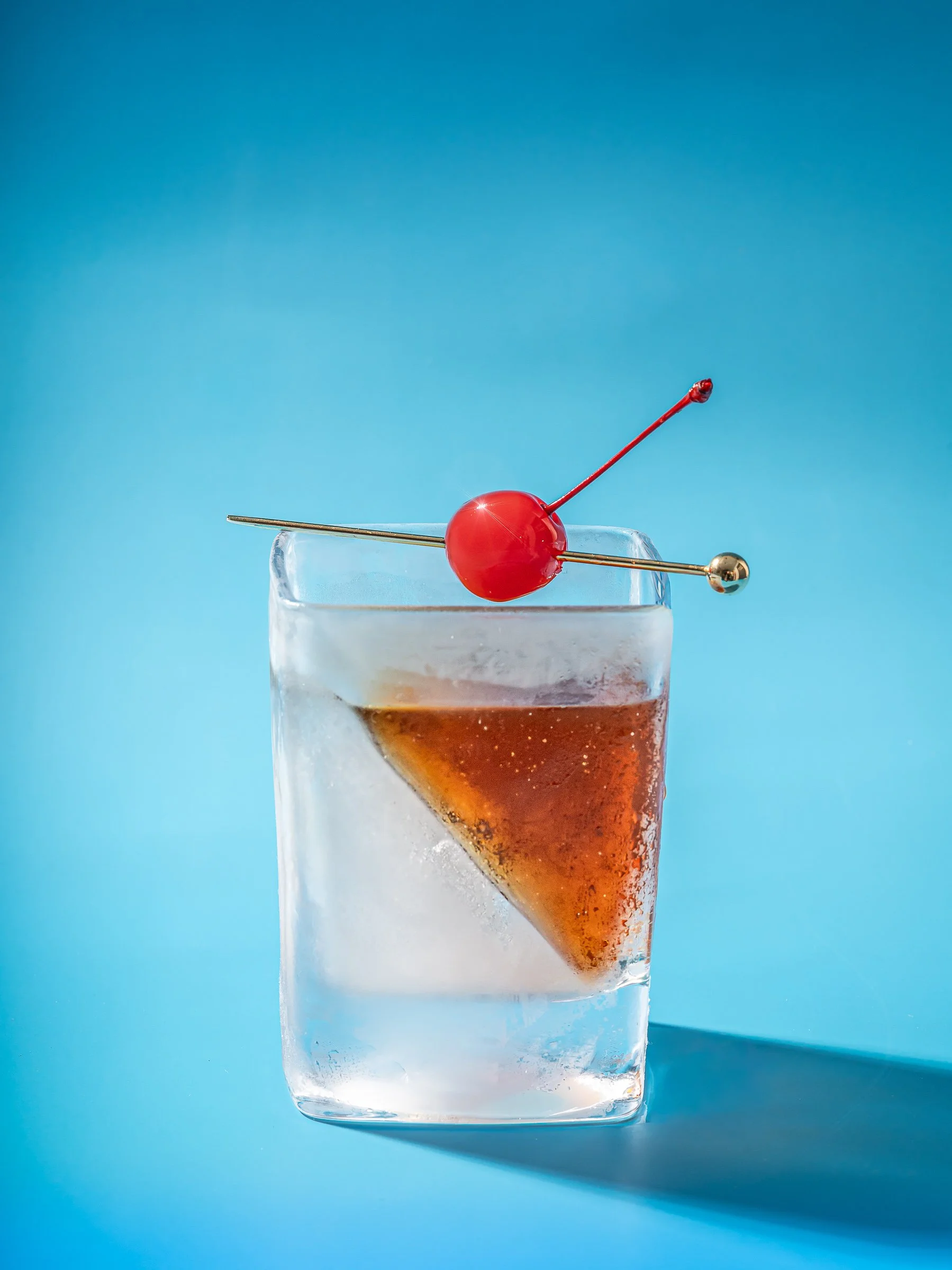 Commercial beverage photography in Miami featuring a whiskey cocktail with large ice and cherry garnish, using bold color lighting for restaurant branding and drink marketing.