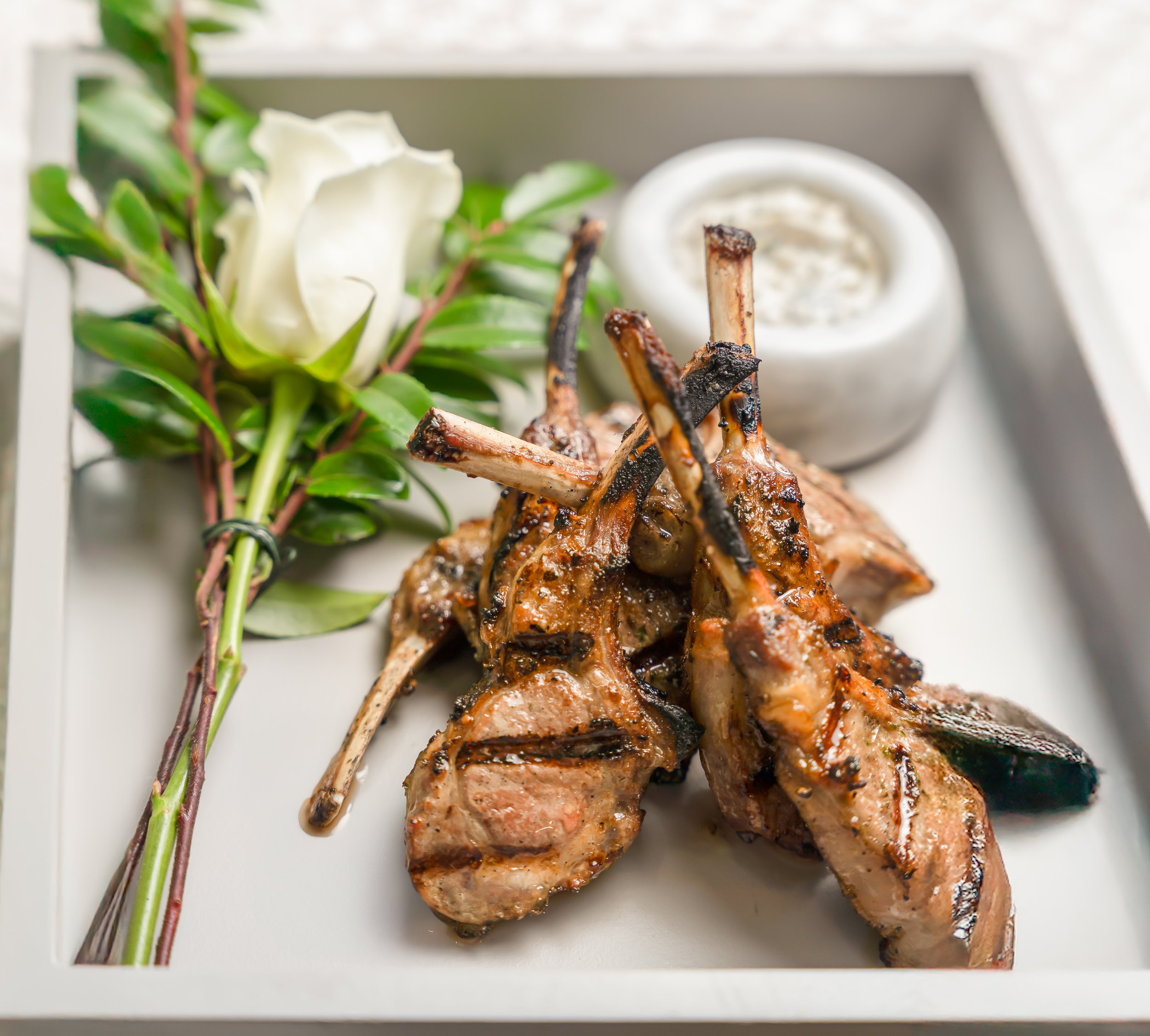 Grilled lamb chops with sprigs of greenery and a white flower, with a small bowl of sauce on a white plate.