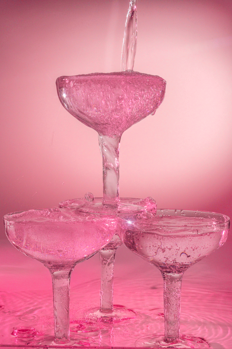 Pink-tinted glasses stacked to form a pyramid with water pouring into the top glass, creating a bubbly splash.