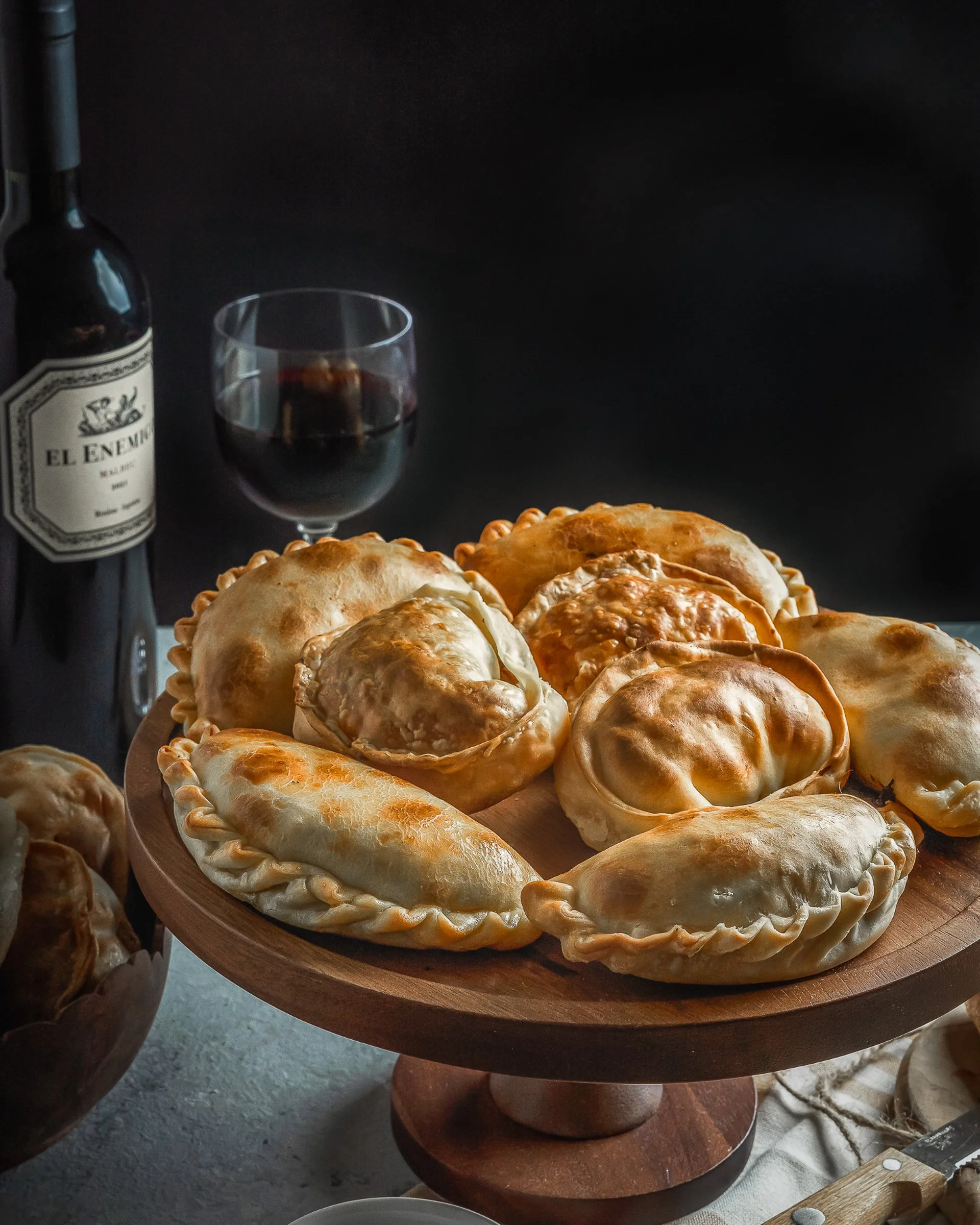 Empanadas Argentinas Tradicionales y Maridaje de Vinos
Fotografía gastronómica editorial de empanadas argentinas artesanales presentadas en un pedestal de madera. La composición incluye un maridaje con vino tinto El Enemigo Malbec sobre un fondo oscu