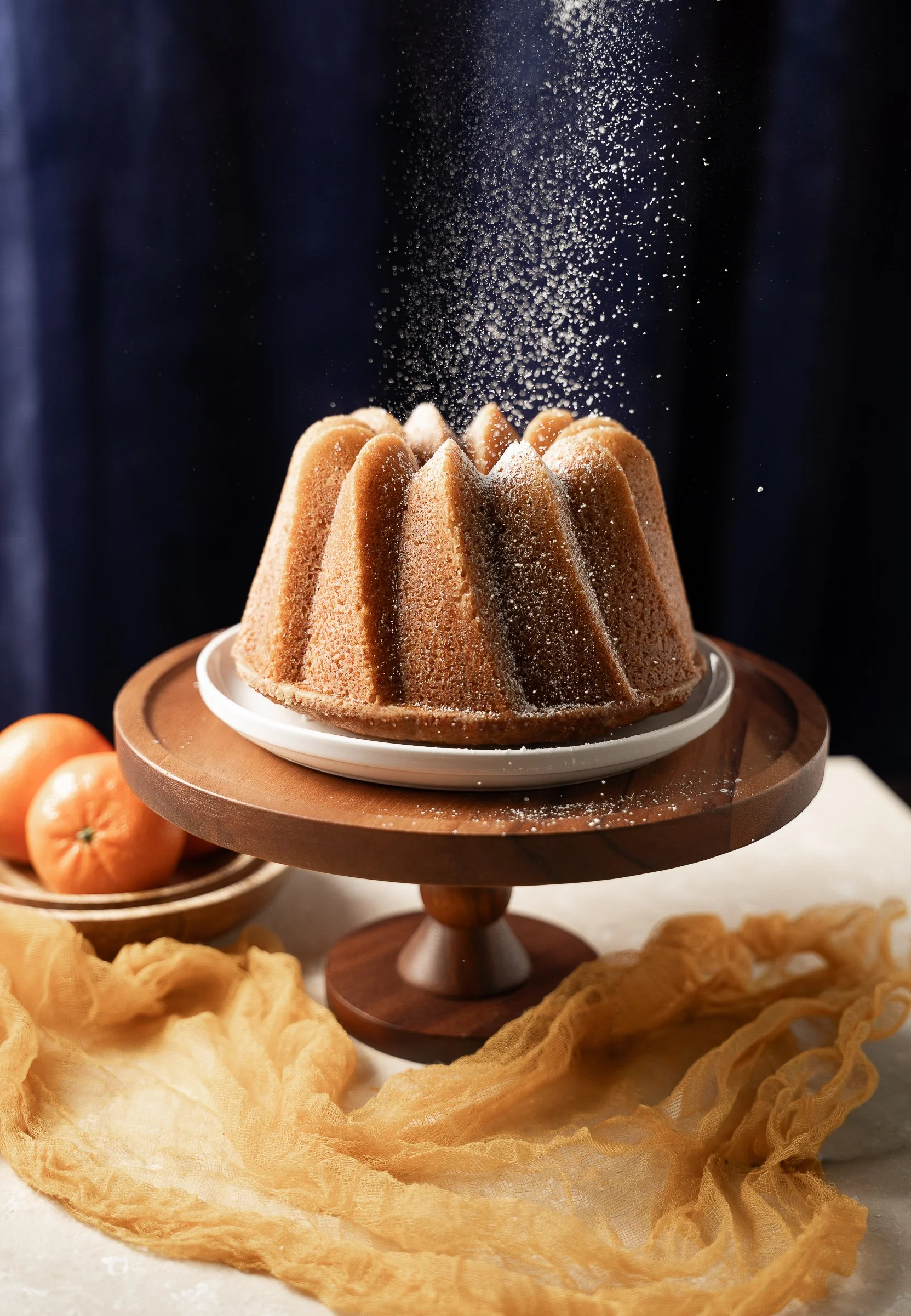 Orange Bundt Cake: Motion & Texture Study
High-speed food photography capturing falling powdered sugar on a fresh orange bundt cake. This editorial shot features warm citrus tones, a wooden pedestal stand, and soft textile textures, demonstrating mas
