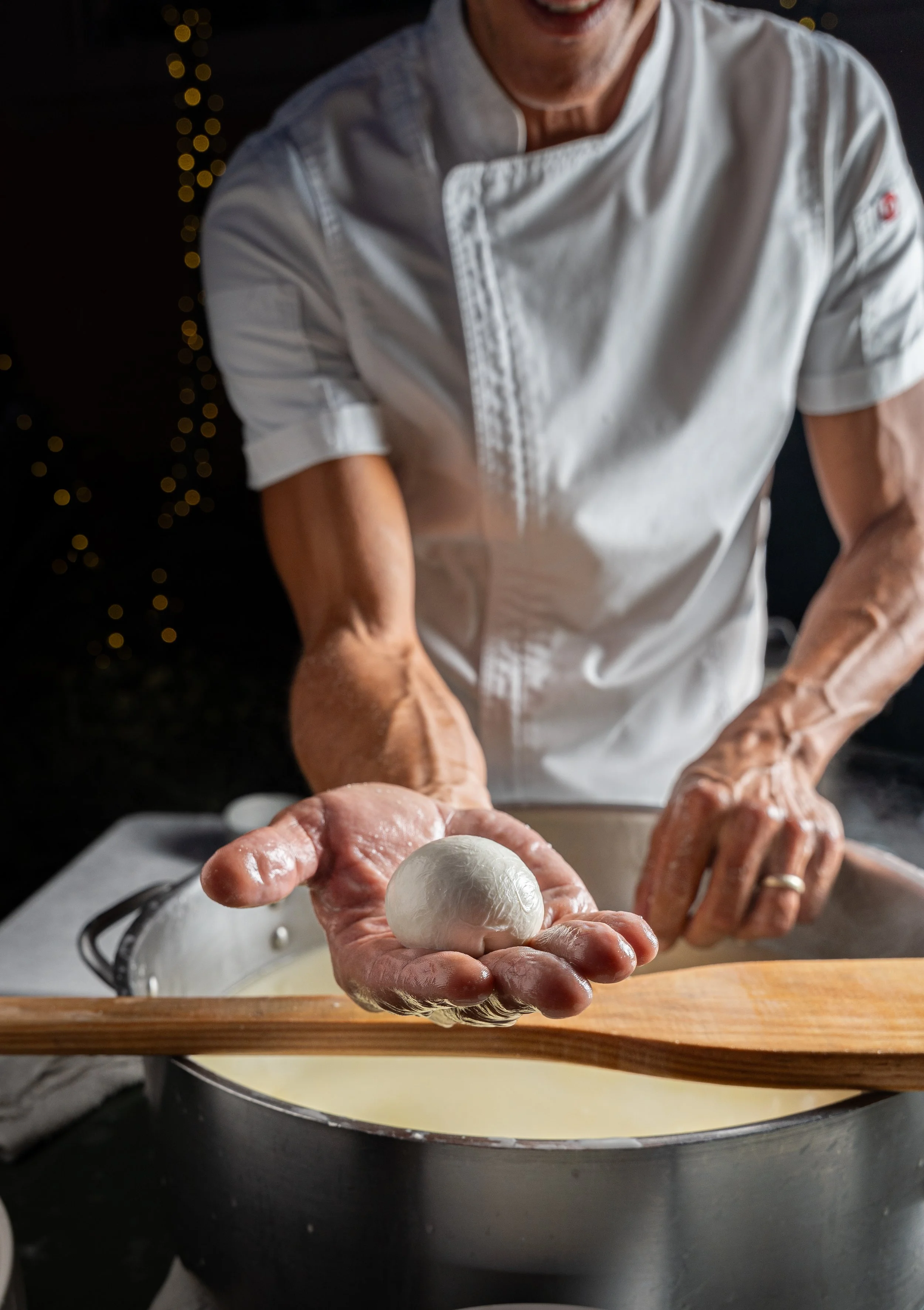 A chef holding a ball of mozzarella cheese over a pot of melted cheese, with a wooden spoon on a marble surface.