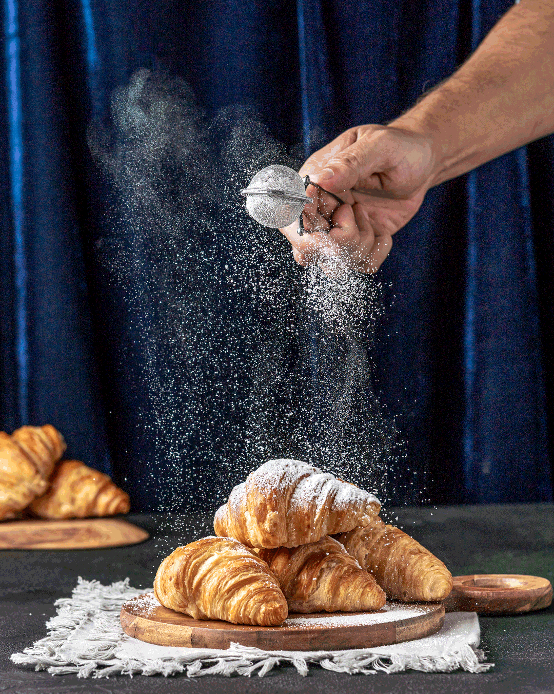 A person dusting powdered sugar onto a plate of croissants with a tea infuser. Croissants are on a wooden board on a cloth, with more croissants in the background.