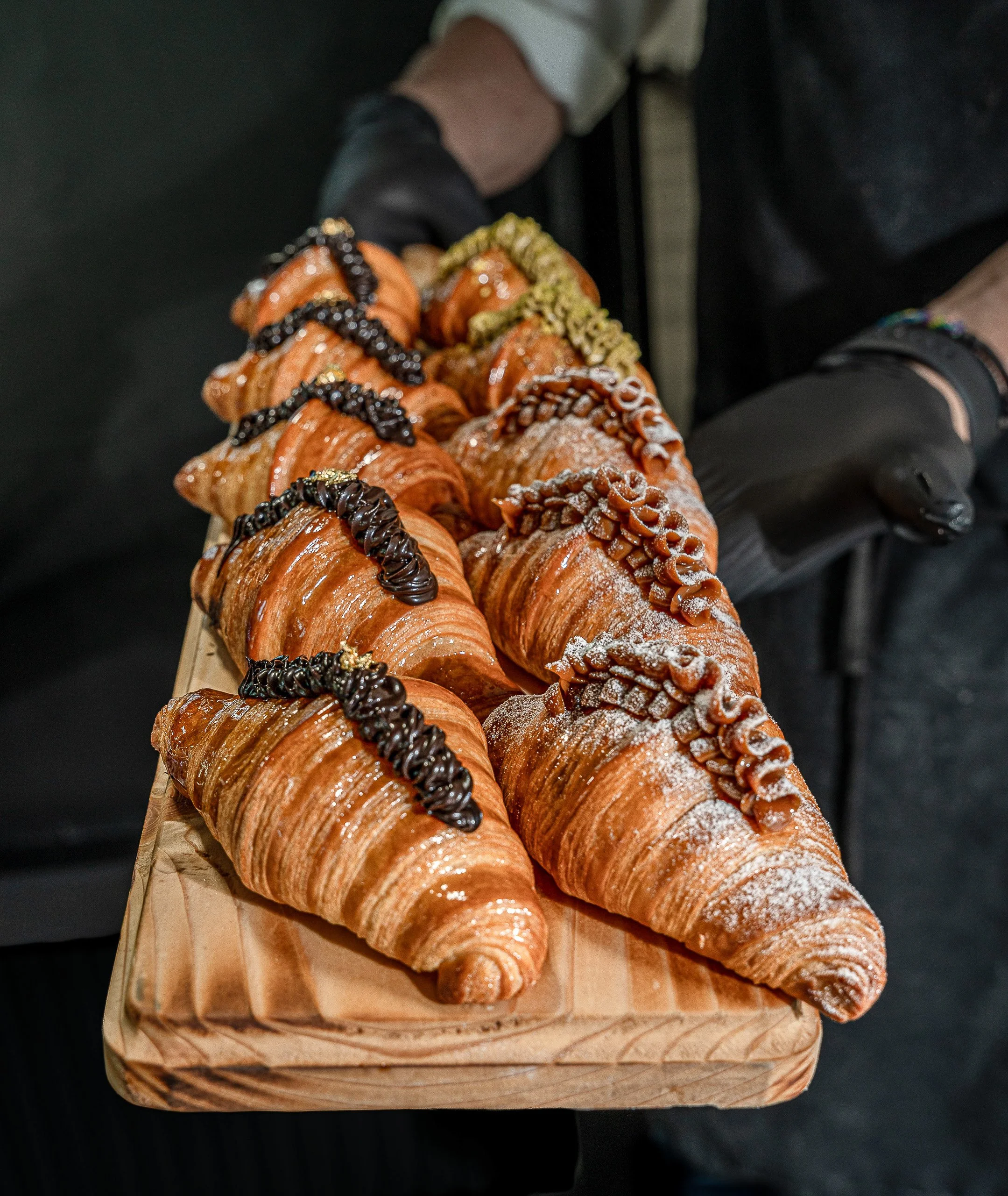 Assorted pastries, including croissants with chocolate drizzle, pistachio toppings, and powdered sugar, arranged on a wooden tray.