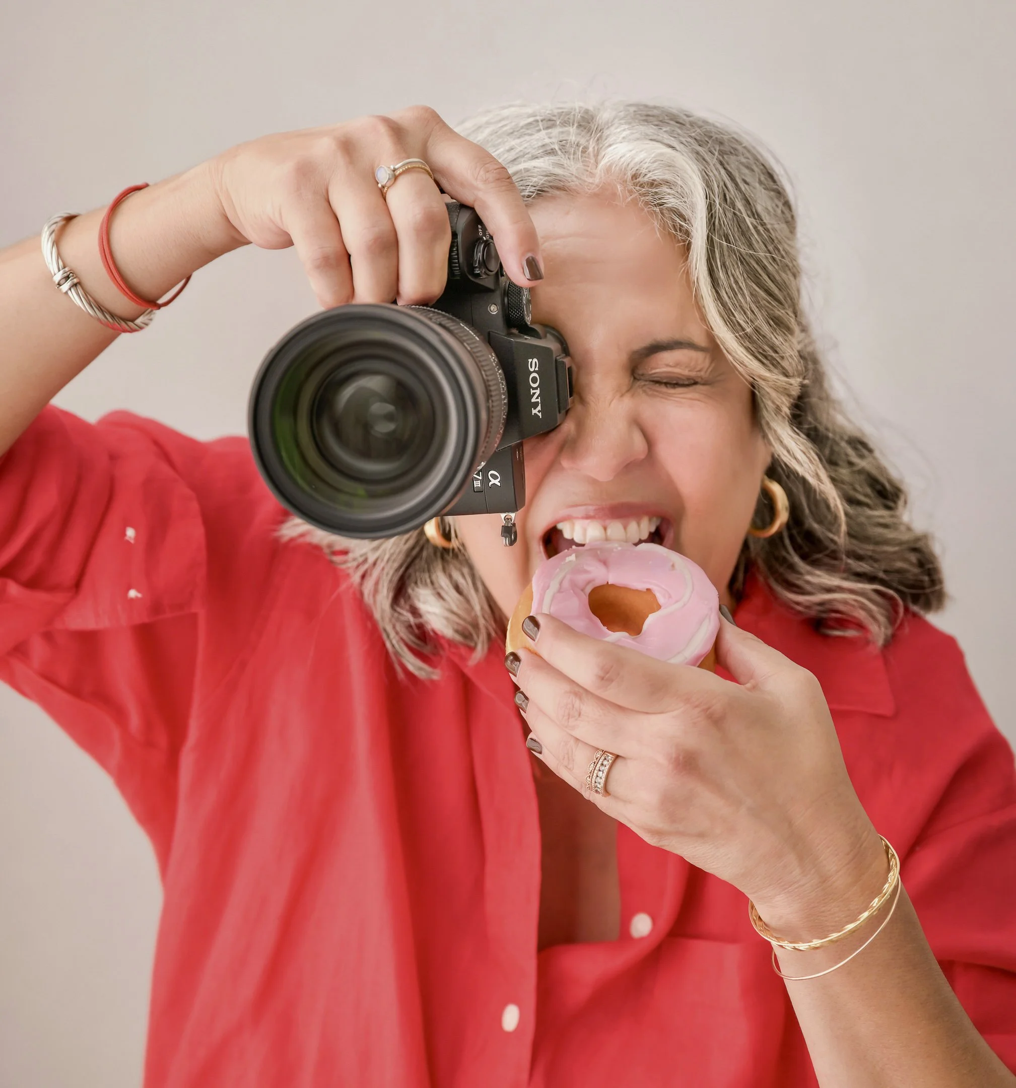 A woman with silver hair wearing a red shirt, jewelry, and rings, is holding a camera and taking a photo while smiling and eating a pink frosted donut with a bite taken out of it.
