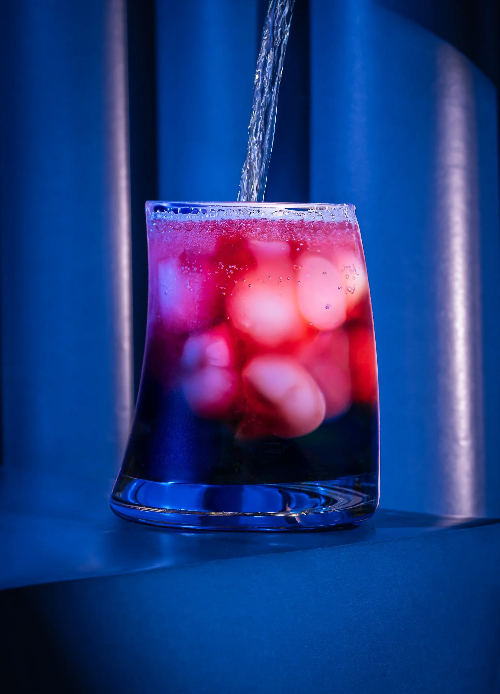 Clear glass containing ice cubes with a pink liquid, with water being poured into it against a dark blue background.