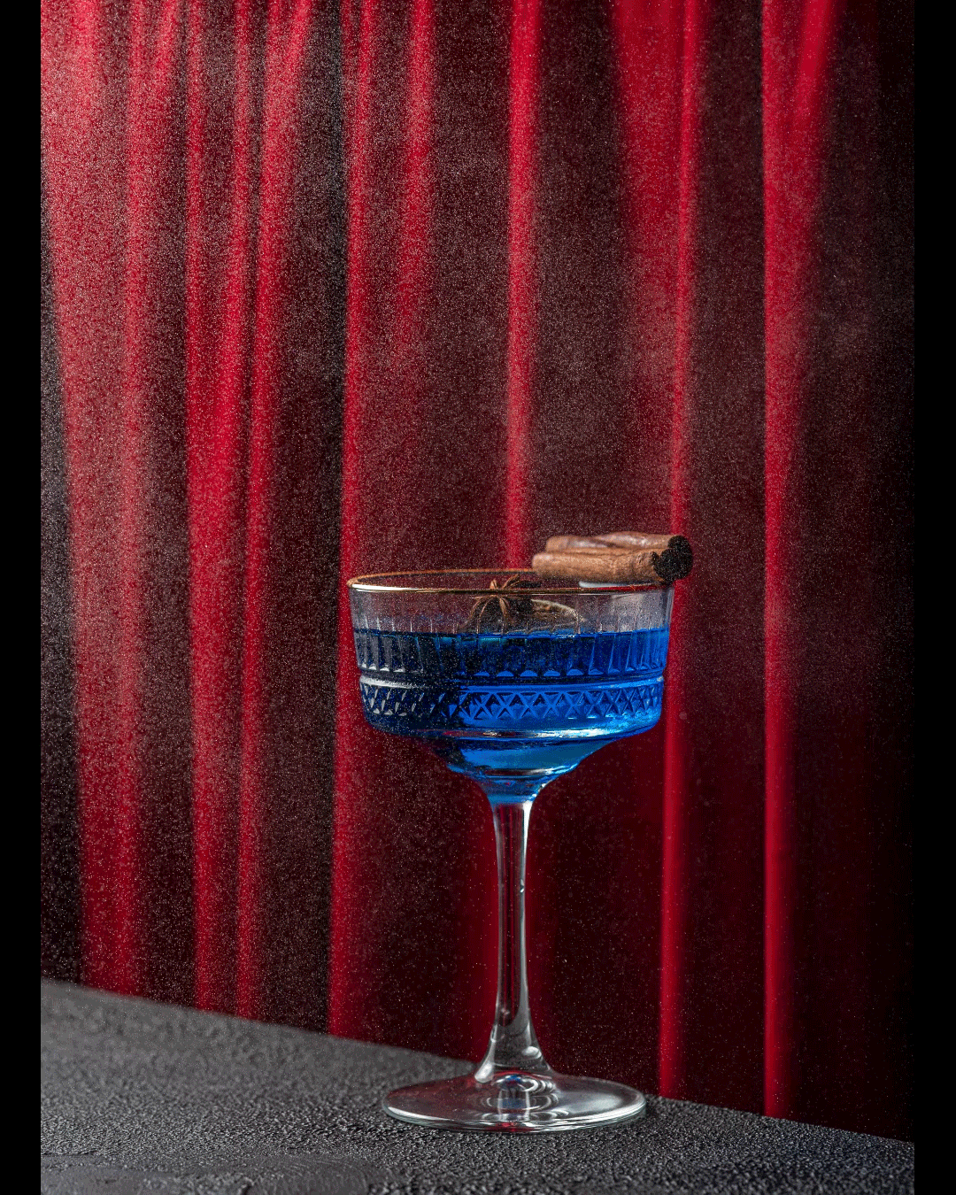 A glass with a blue cocktail garnished with a piece of cinnamon stick, set against a red curtain background.