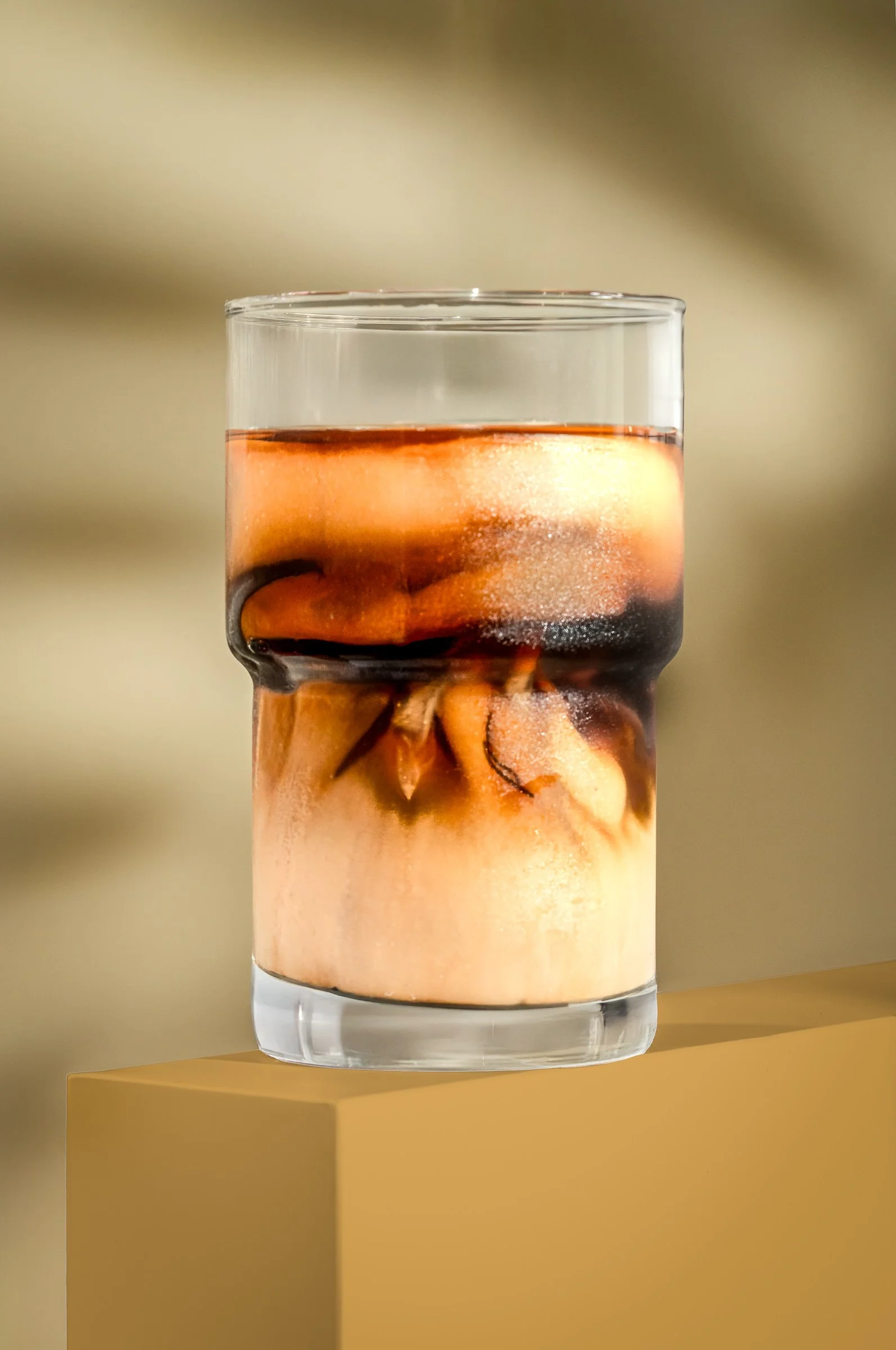 Close-up of a glass of iced coffee with cream, ice cubes, and coffee swirling together, placed on a yellow surface.
