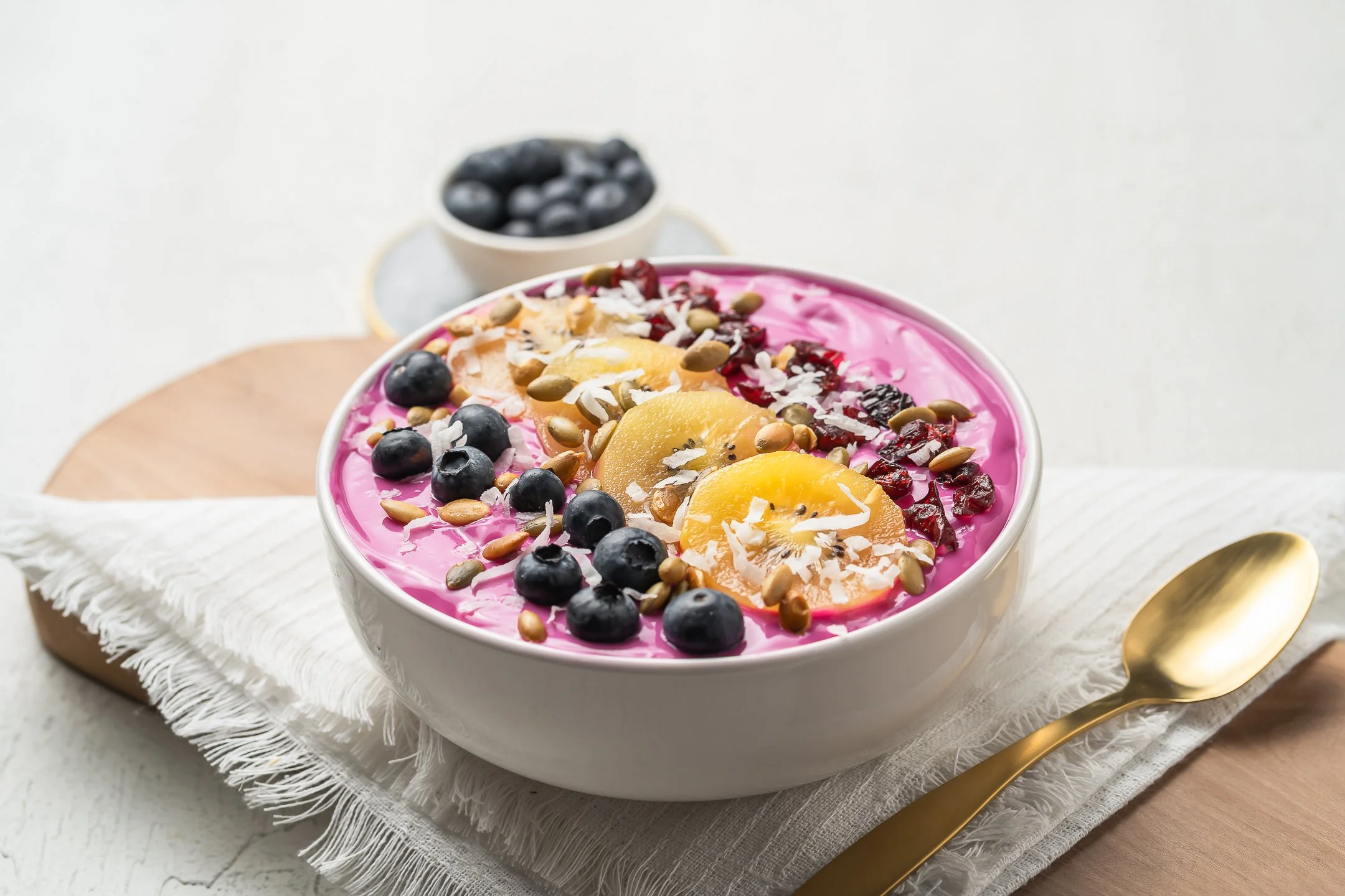 A bowl of pink yogurt topped with blueberries, peach slices, dried cranberries, shredded coconut, and pumpkin seeds, placed on a white cloth with a gold spoon beside it, on a wooden board.