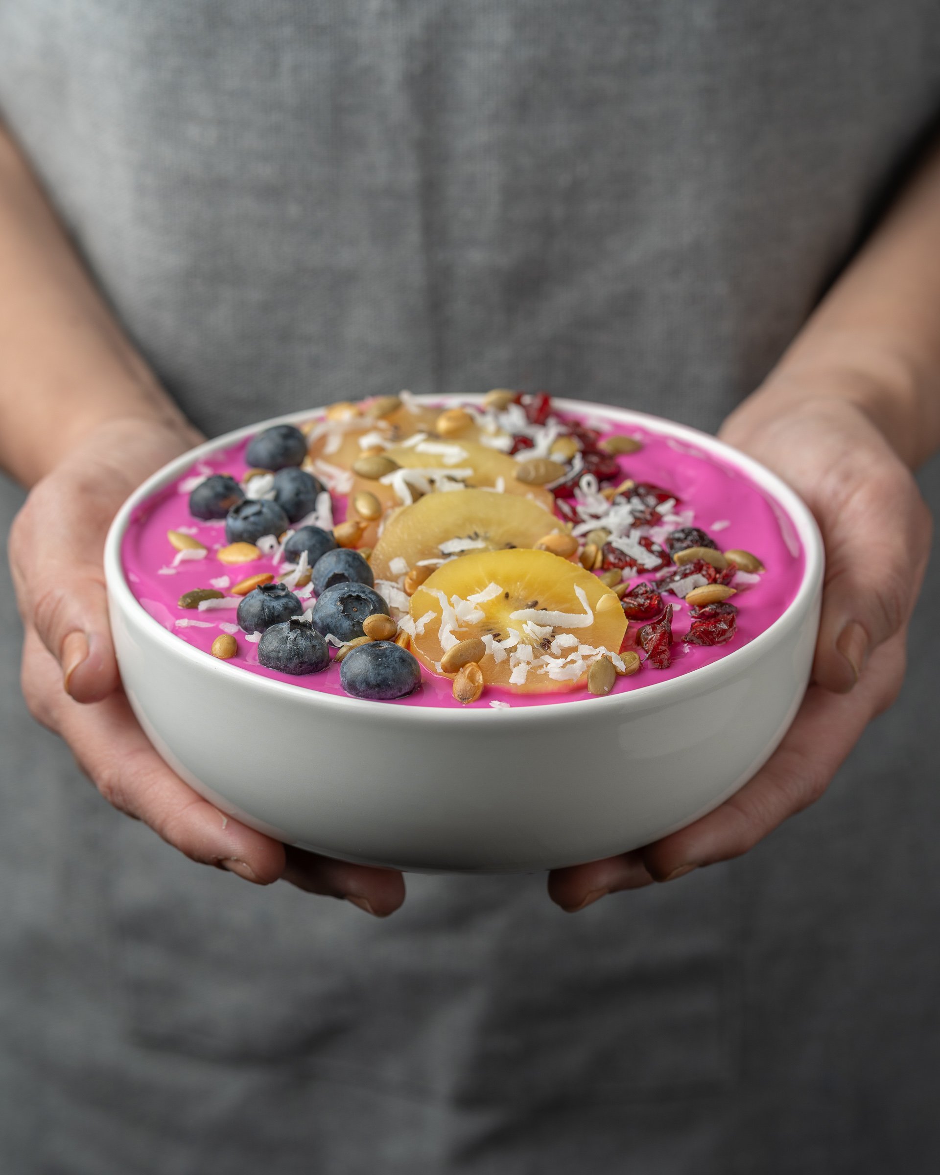 Vibrant Pitaya Bowl | Lifestyle Food Photography
A bright, colorful lifestyle food photograph of a dragon fruit smoothie bowl held by a person in a grey apron. The bowl is meticulously styled with blueberries, golden kiwi, coconut, and seeds, showcas