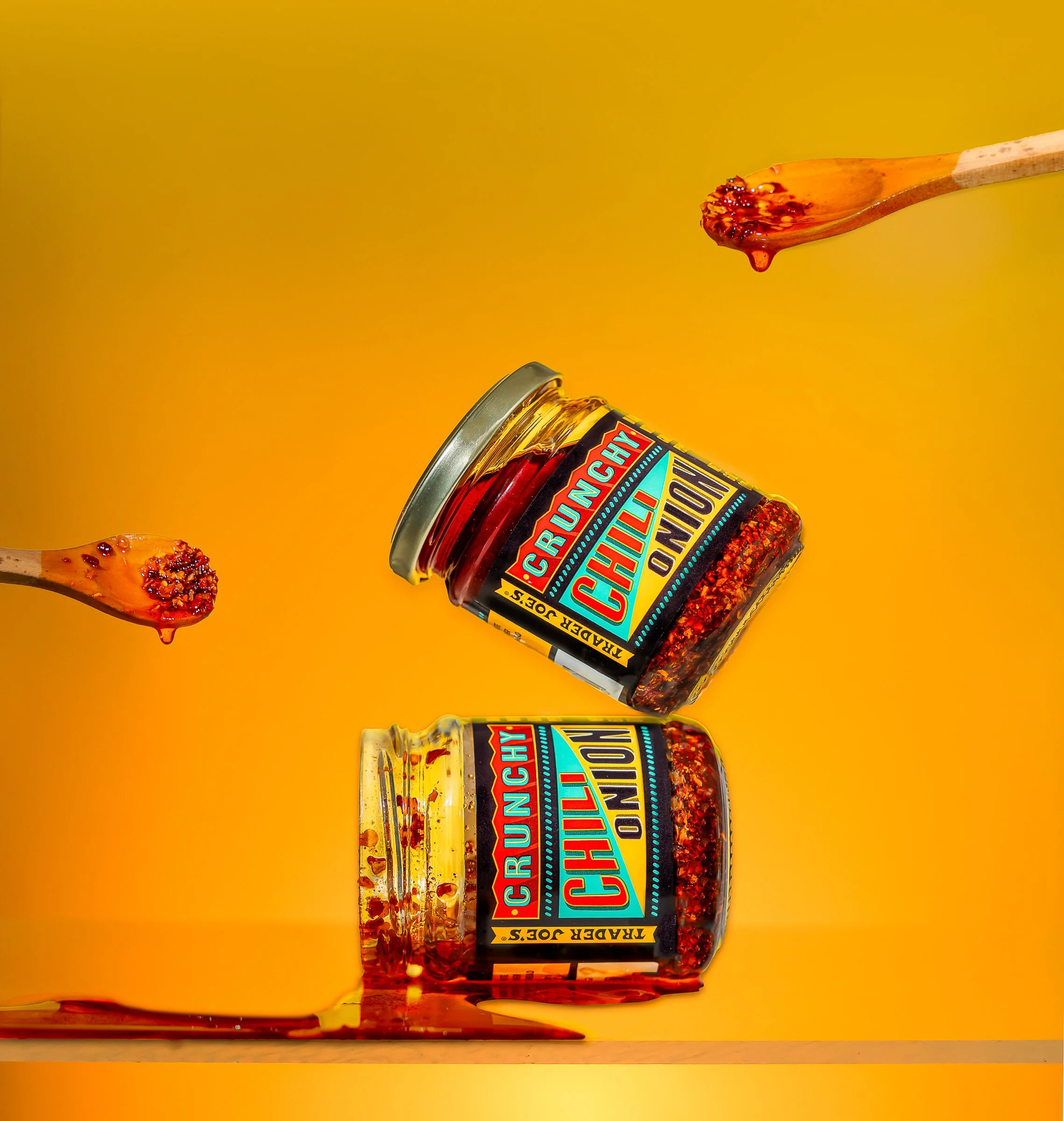 A jar of spicy chili flakes labeled 'Honey Pecan & Chili' with some chili flakes spilled on the surface and a wooden spoon with chili flakes dripping from it, set against a yellow background.