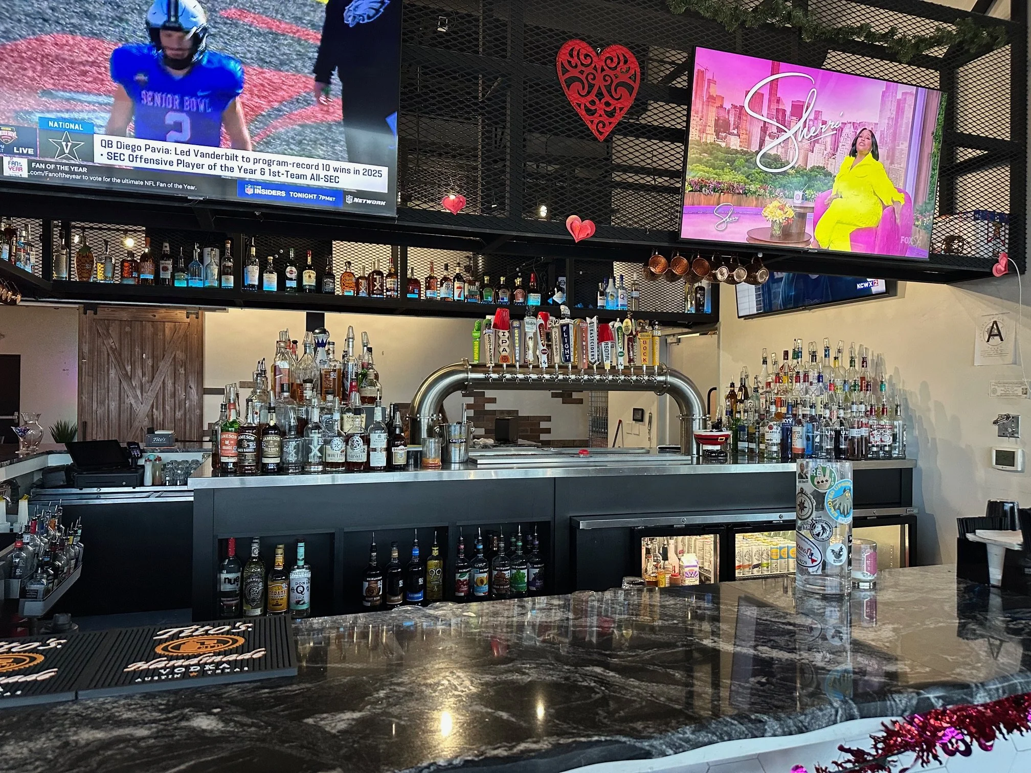 Bar area with various liquor bottles on shelves, two televisions displaying different programs, heart-shaped decorations hanging, and a floral arrangement on the counter.
