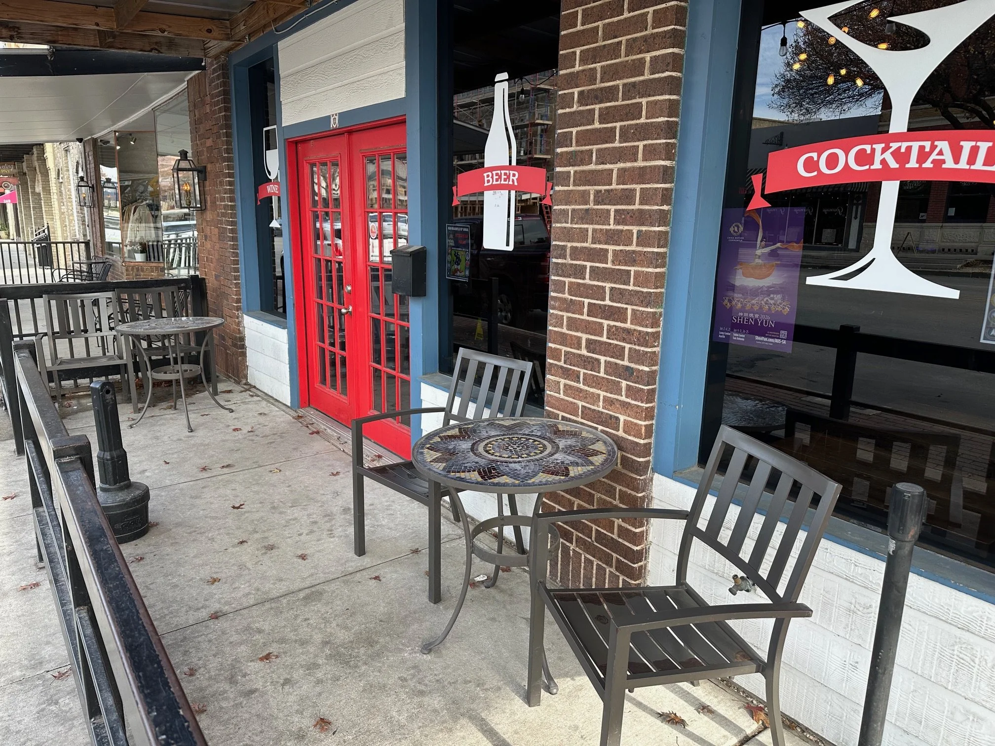 Outdoor seating area of a restaurant with metal chairs and small round tables, brick and white siding exterior, red door, and windows decorated with signs indicating wine, beer, and cocktails. There are hanging lantern-style lights and string lights 