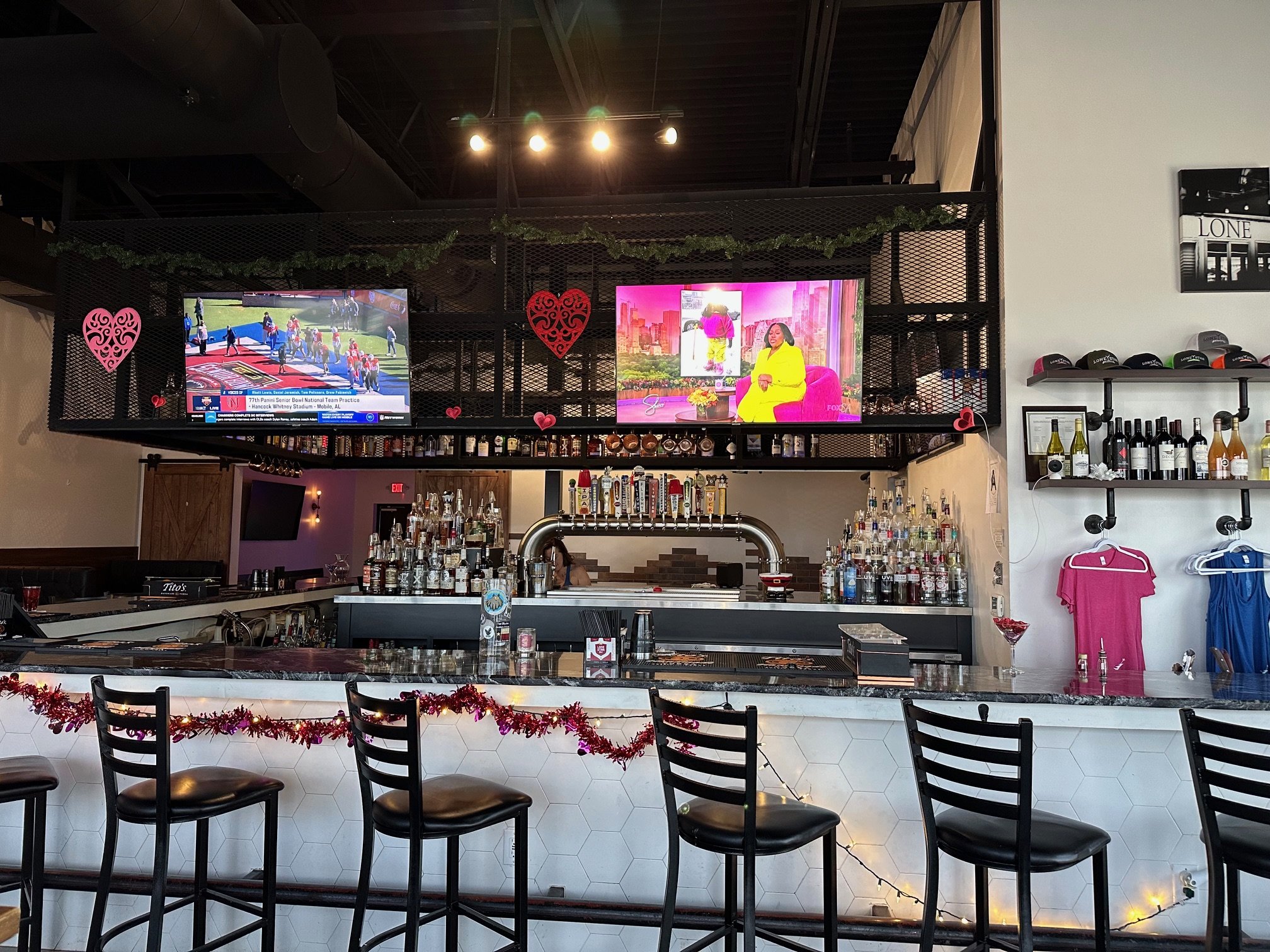 A bar with a marble countertop, black chairs, and Christmas decorations including red garlands. Behind the bar are bottles of alcohol, plus two television screens showing a sports game and a television show. The wall is decorated with black and white