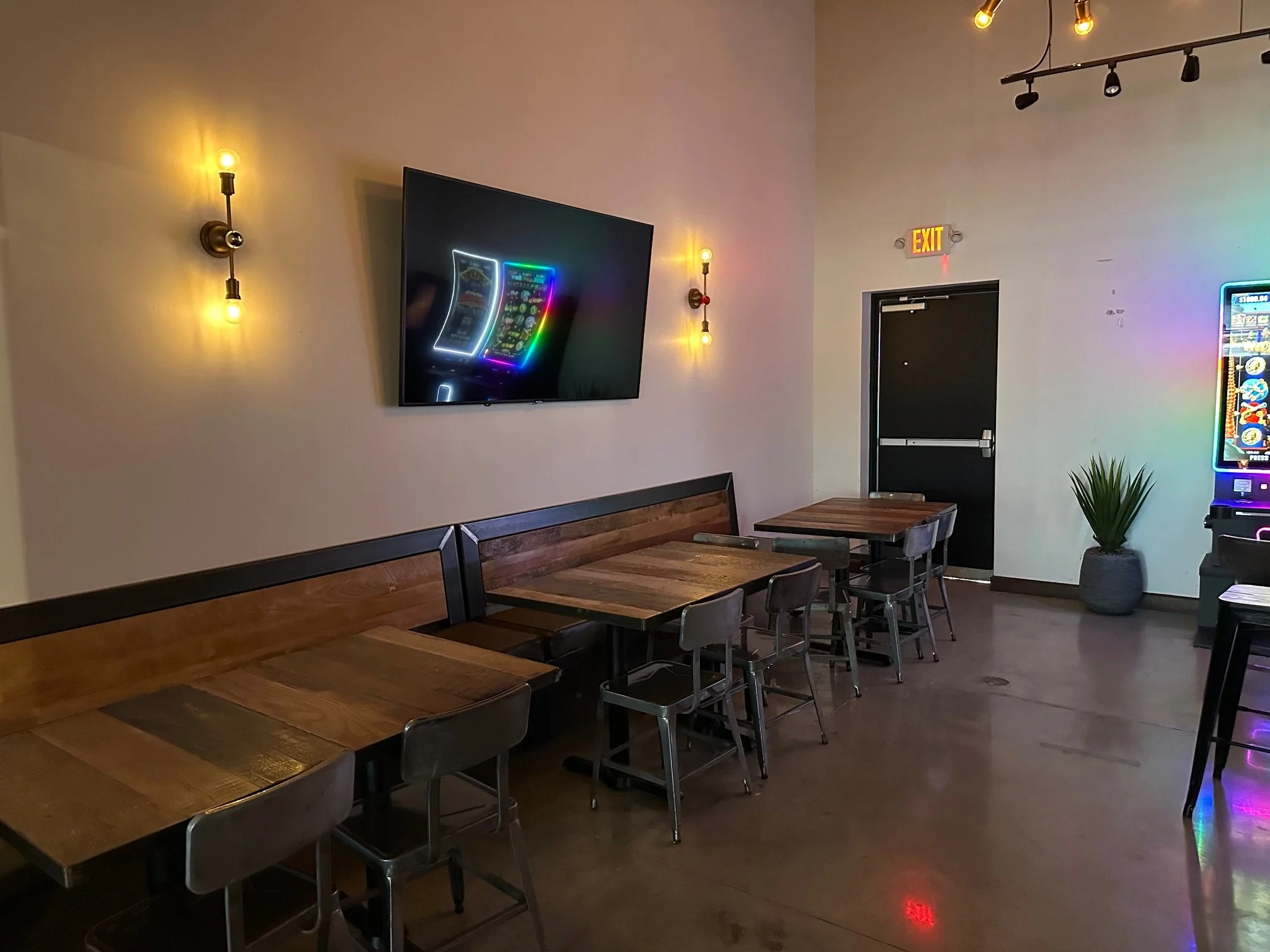 Interior of a restaurant with wooden tables and gray chairs, decorated with wall-mounted lights, a flat-screen TV on the wall, and a plant in a pot near a colorful arcade game machine. There is an exit door with an illuminated red sign above it.