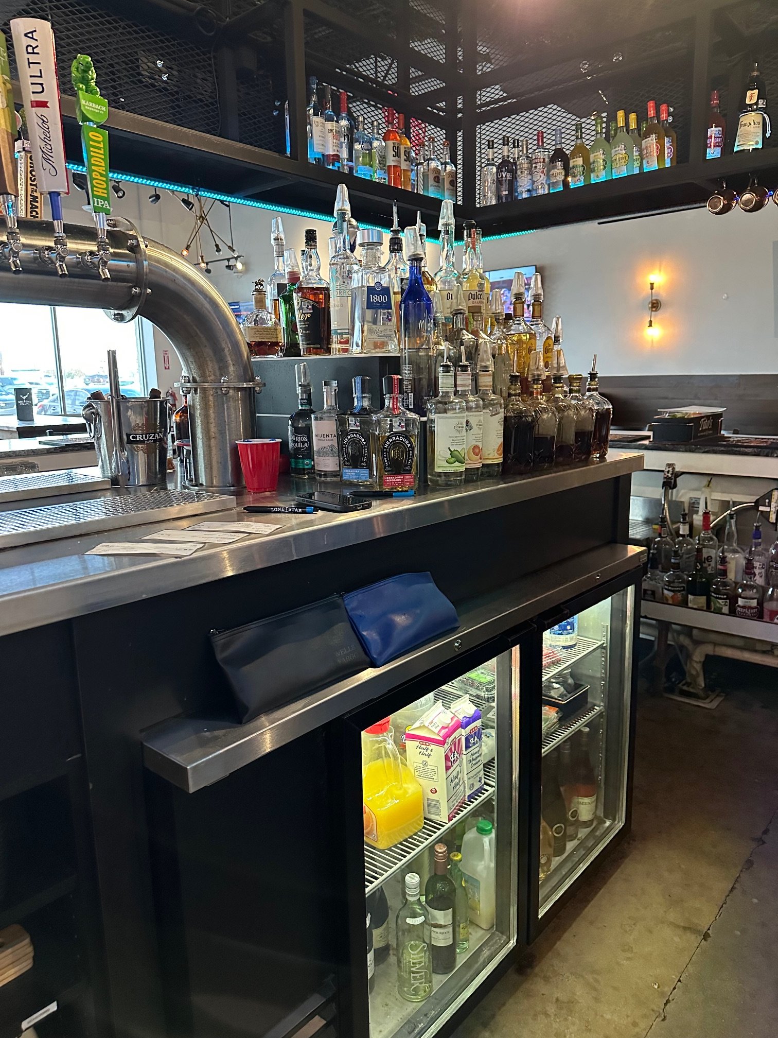 A bar with various bottles of liquor on shelves, a beer tap system, a red cup, and a refrigerator with beverages inside.