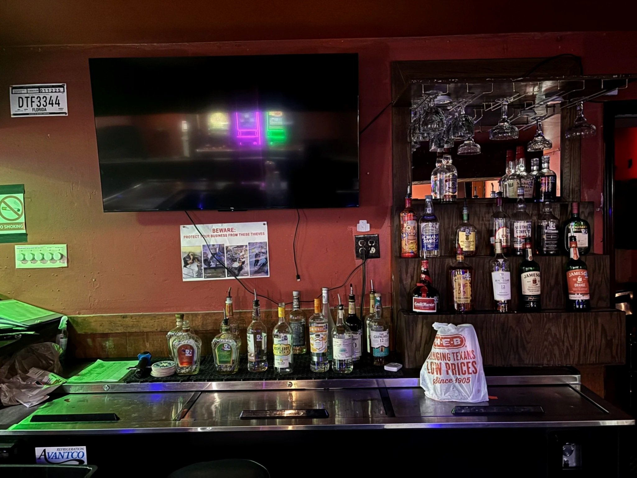 Bar area with liquor bottles, hanging glassware, a television screen, and various signs, including a no smoking sign and a warning about protection from thieves.