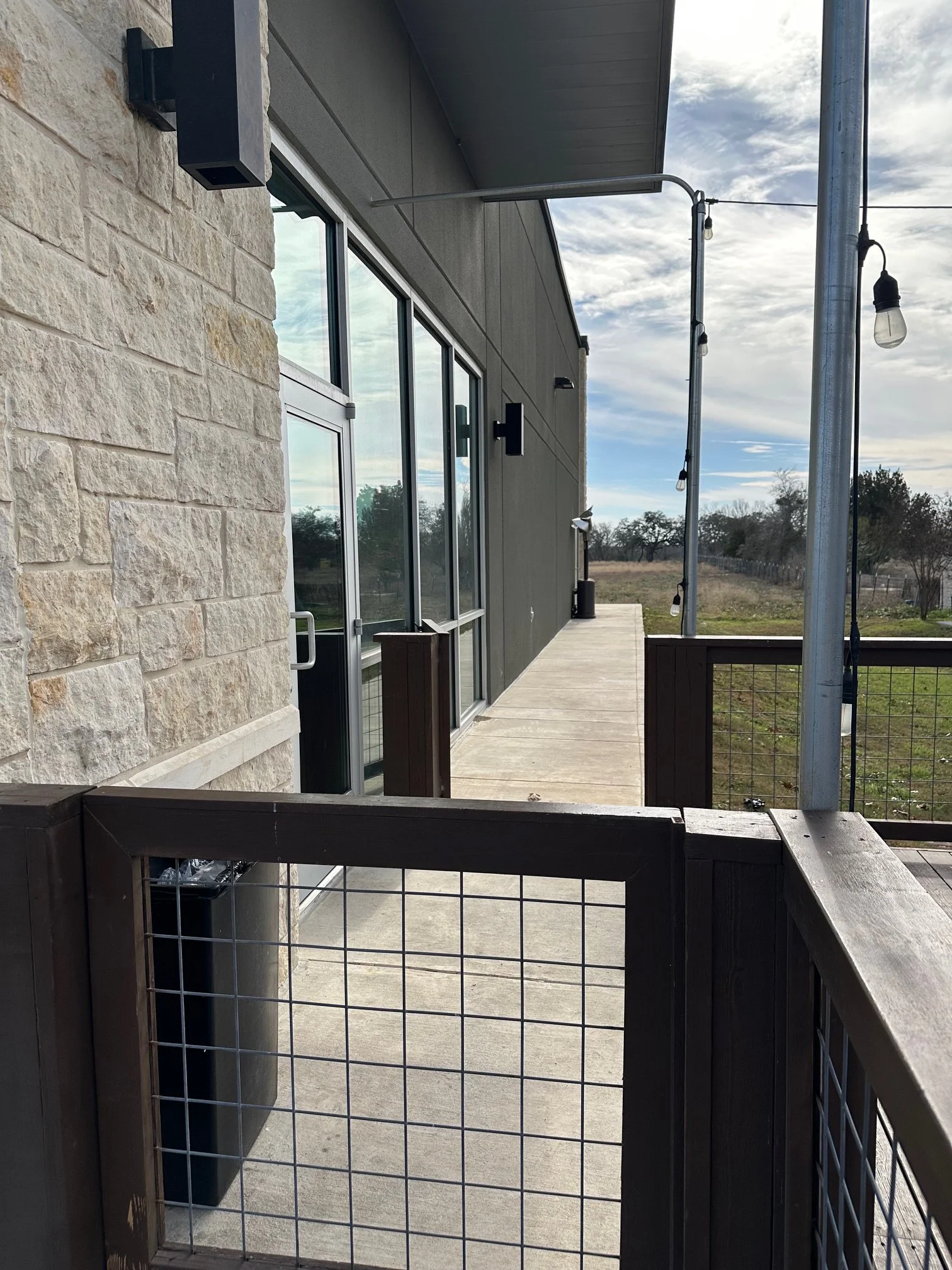 A photo of a balcony area outside a modern building with stone and gray panel walls. It features large glass doors, an outdoor string light, and a railing with a metal wire grid. In the background, trees and a partly cloudy sky are visible.