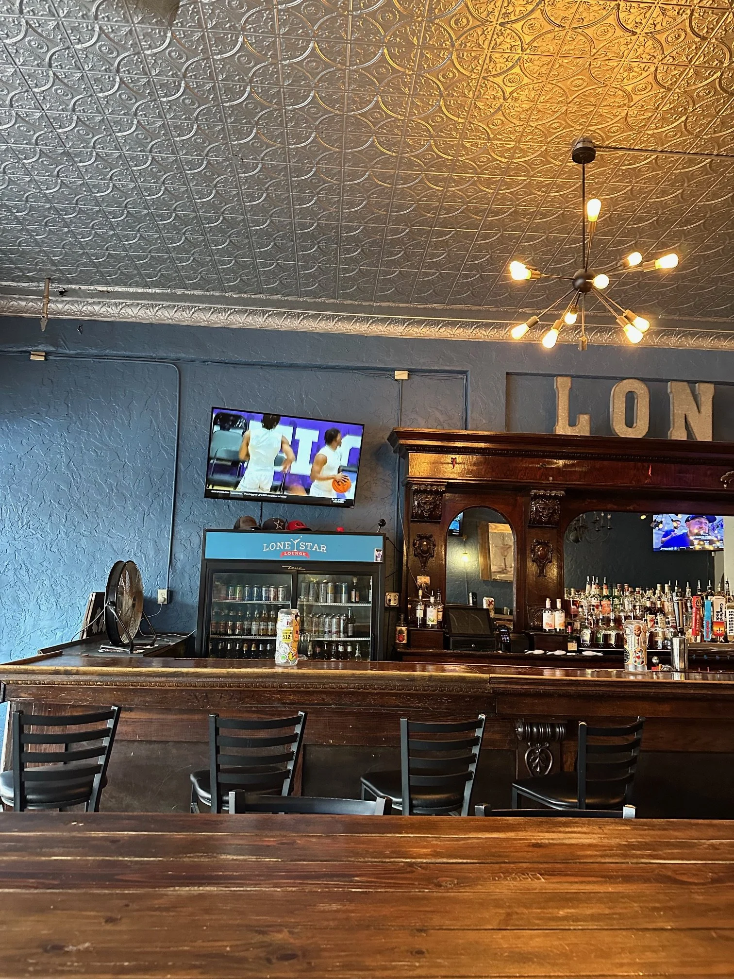 Interior of a bar with a wooden counter, black chairs, a blue textured wall, TV screens showing sports, and a shelf with bottles. The ceiling has intricate metallic tiles and a modern chandelier with exposed bulbs. The word 'LON' is partially visible