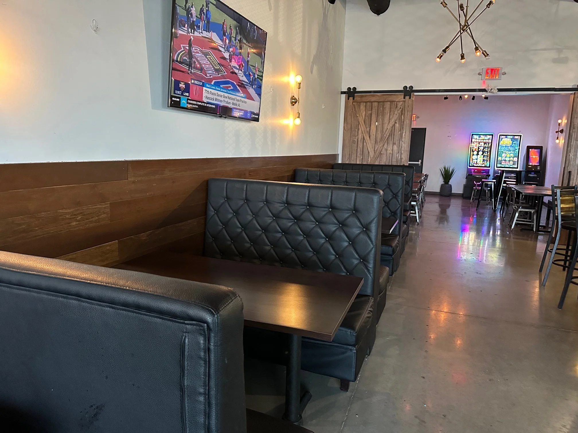 The image shows a modern lounge or game room with a large flat-screen TV mounted on the wall, playing a sports game. There are dark leather booths and tables in the foreground and additional seating with arcade game machines in the background. The ro