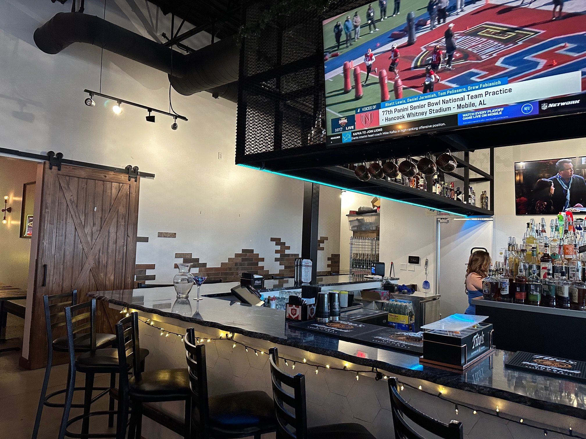 Interior of a sports bar with a large screen showing a football practice, a bar counter with liquor bottles, a bartender, and seating area with chairs and a table decorated with string lights.