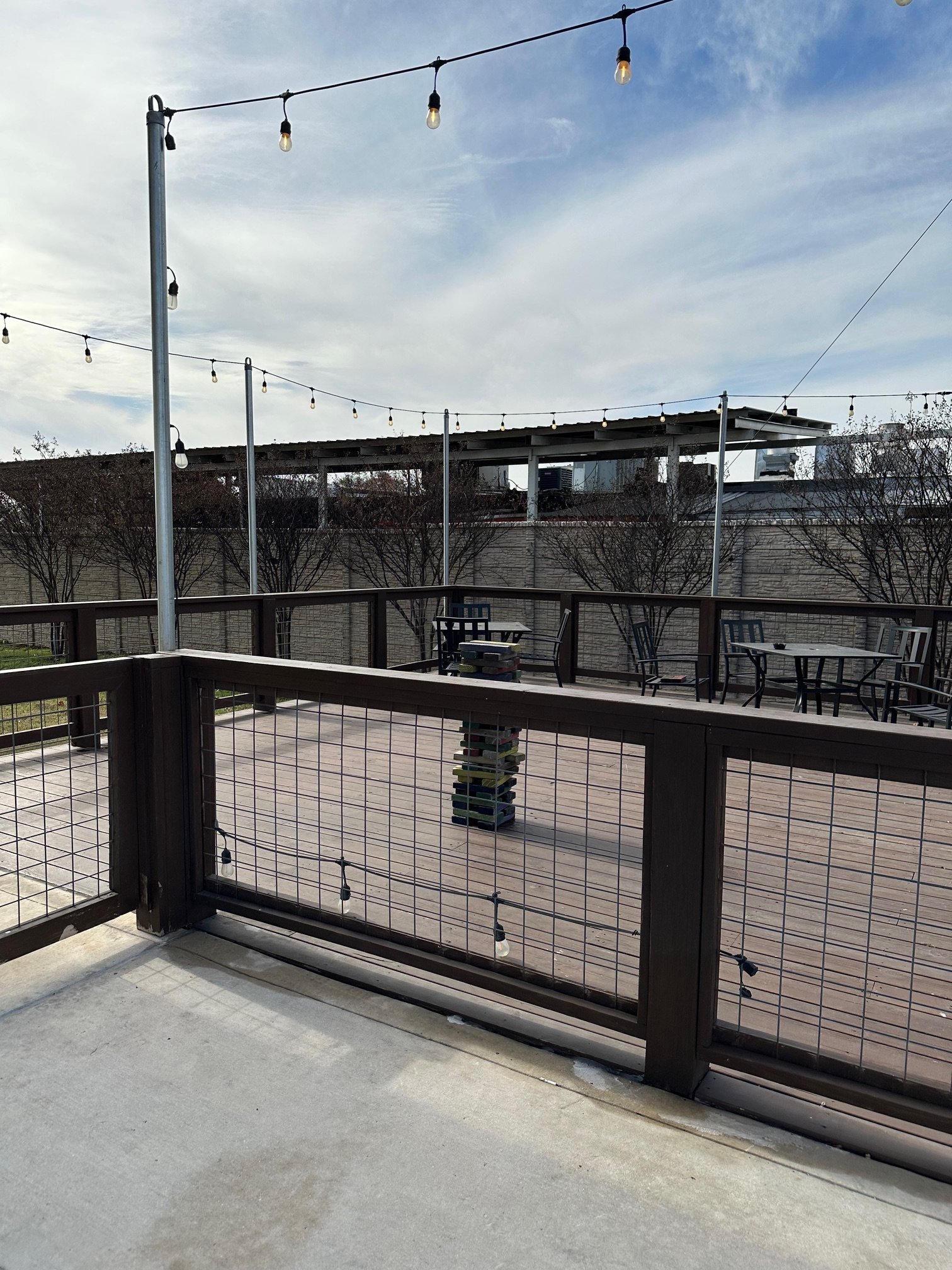 An outdoor patio area with a wooden railing and string lights. There are several patio chairs and a small stack of books on a table. A fence surrounds the area, and there are bare trees in the background. The sky is partly cloudy.