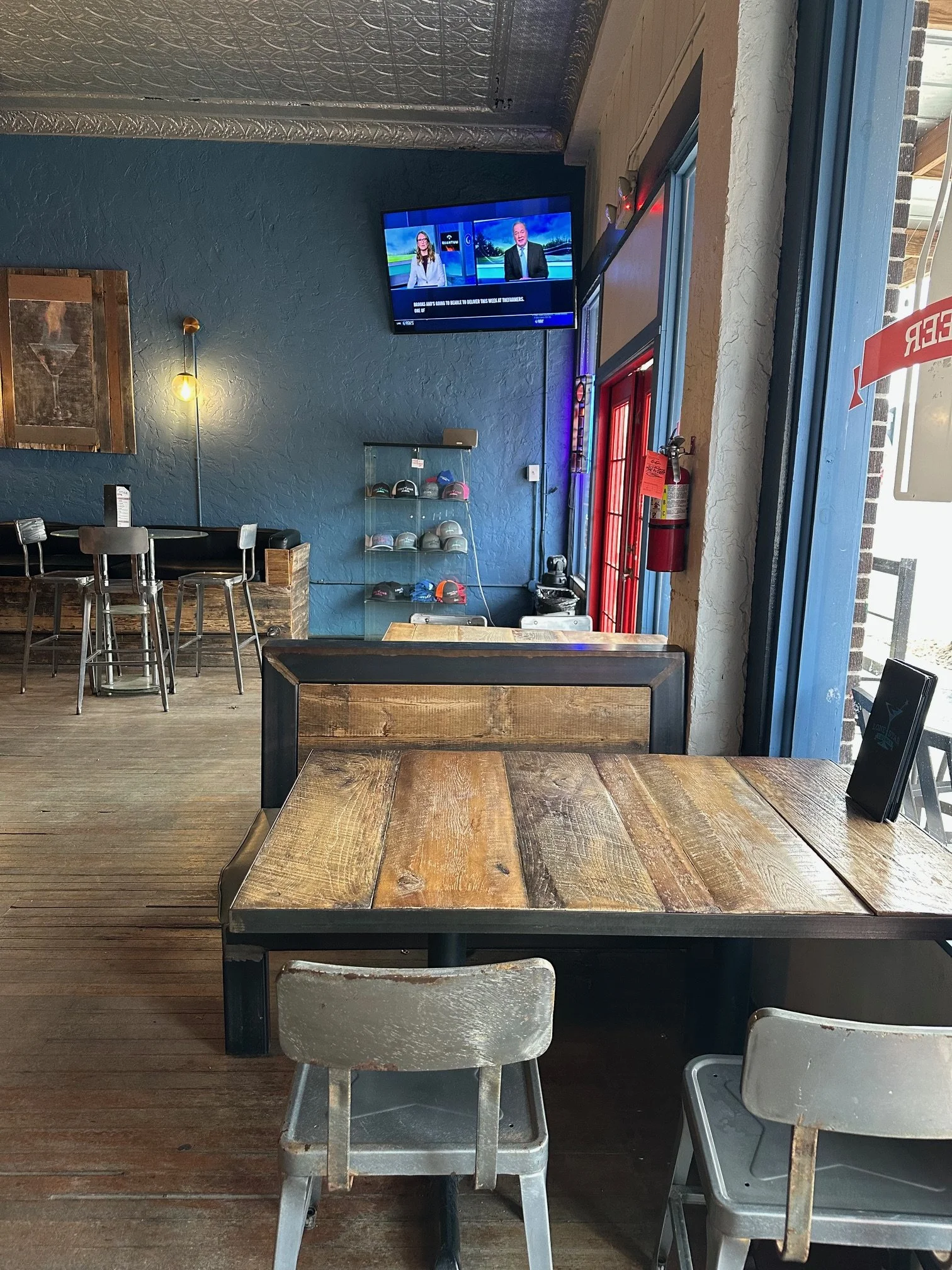 A rustic wooden table with seating in a casual restaurant. The background features a blue wall with a mounted flat-screen TV showing news anchors, and a glass display case with caps. There is a red door or window frame with a fire extinguisher nearby