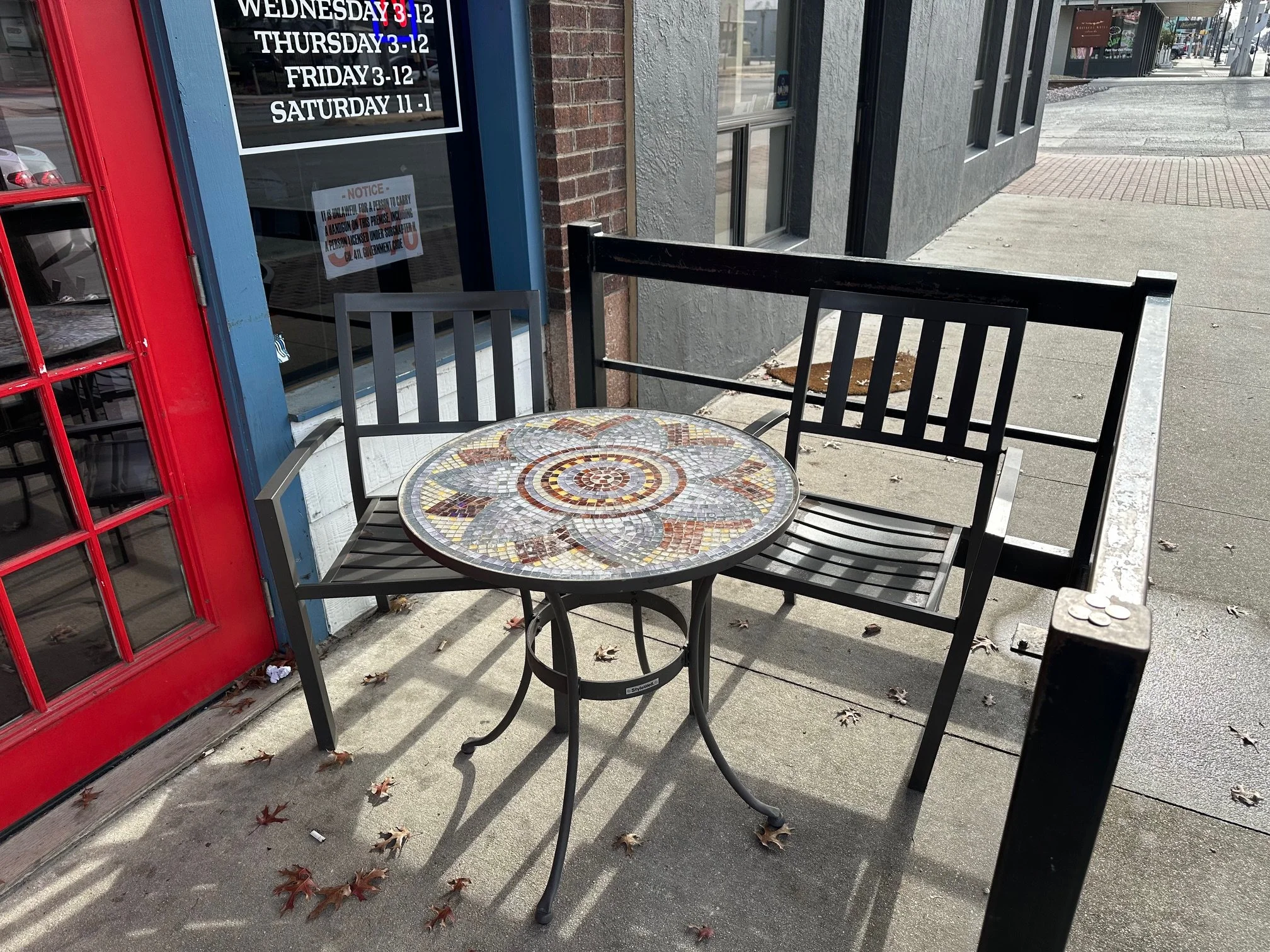 Outdoor patio seating area with a mosaic table and black chairs outside a storefront with a glass door and large window, displaying business hours.