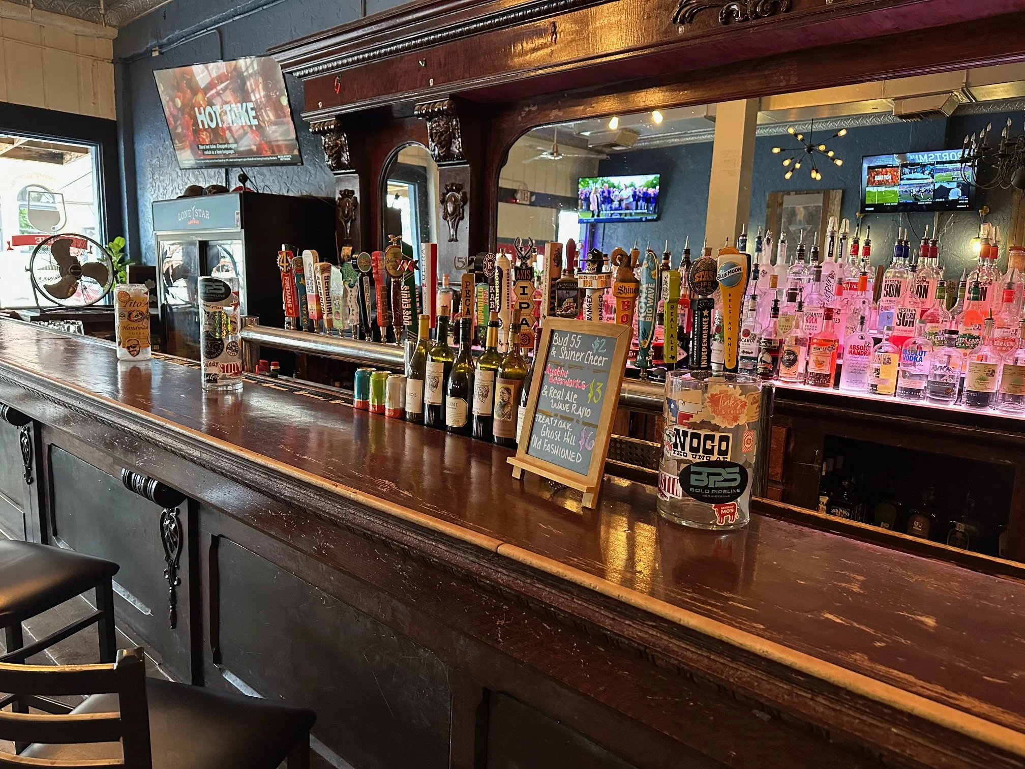 Inside a bar with a row of colorful tap handles, a chalkboard menu, a wine bottle, and TV screens on the wall. The bar has ornate woodwork and a dark color scheme.