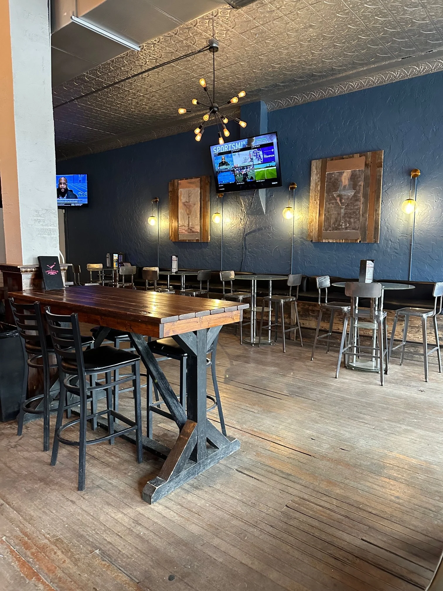 Interior of a bar or restaurant with wooden floor, tables, and chairs, multiple TVs on blue walls showing sports content, modern ceiling light fixture, and wall art.