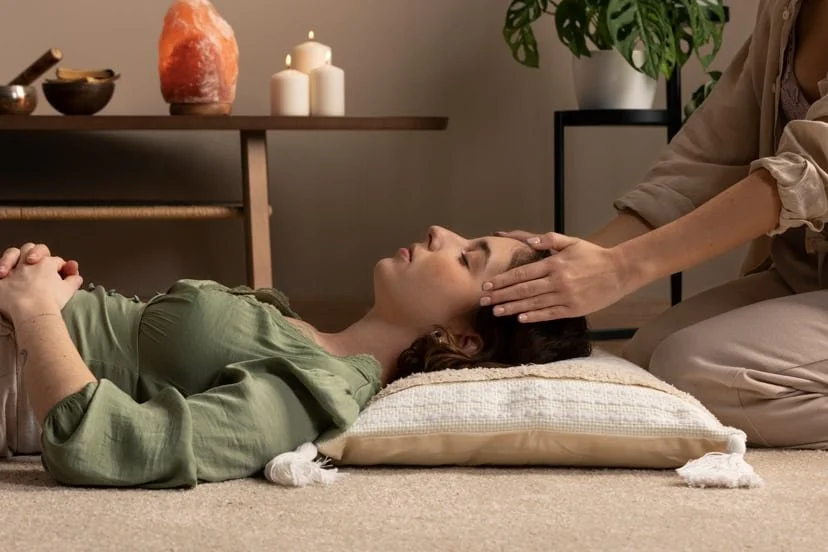 A woman lying on her back with eyes closed, receiving a head massage from a therapist in a cozy room with candles and a salt lamp on a shelf.